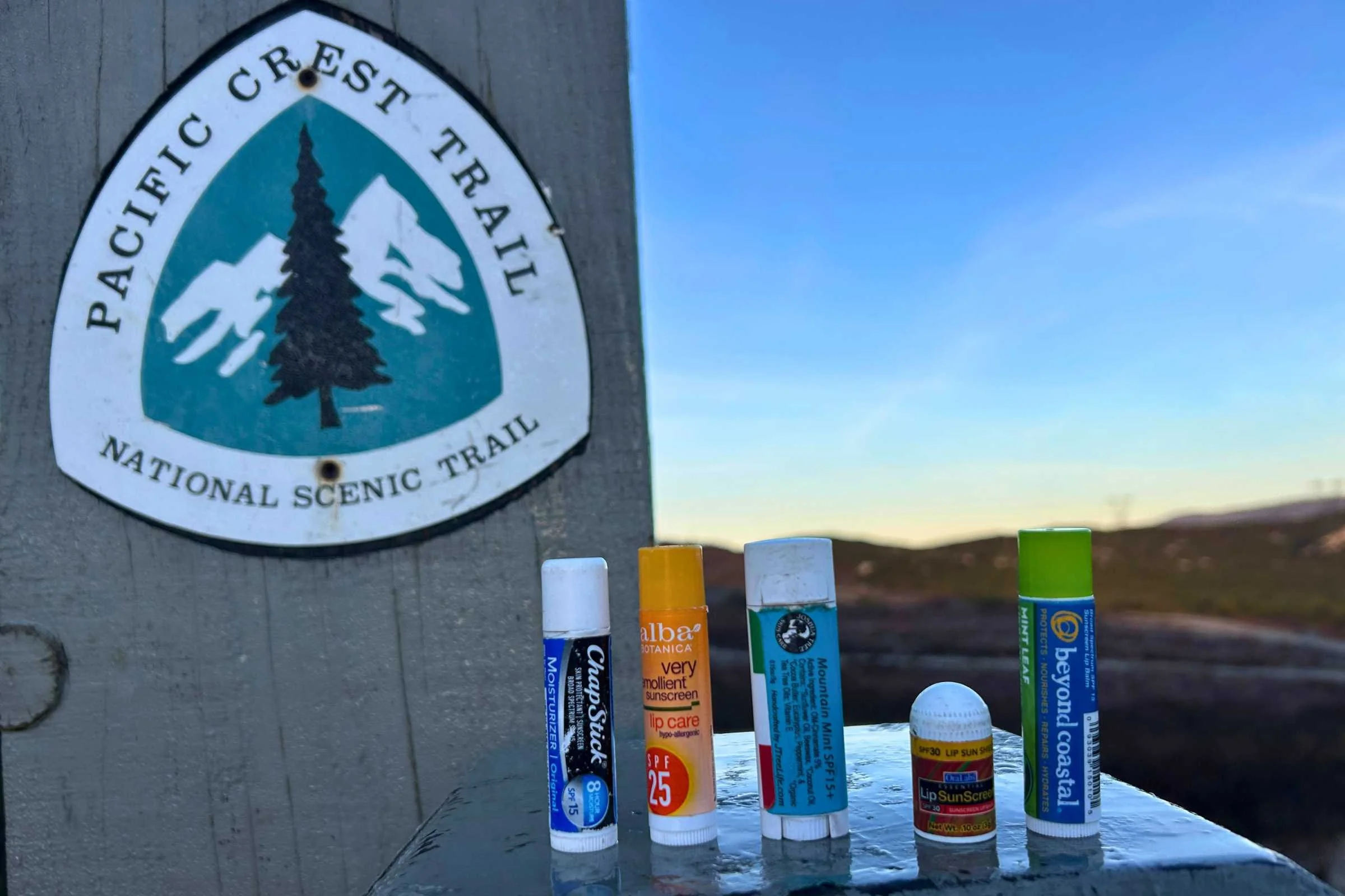 Collection of SPF lip balms displayed beside Pacific Crest Trail sign during outdoor hiking and sun protection testing