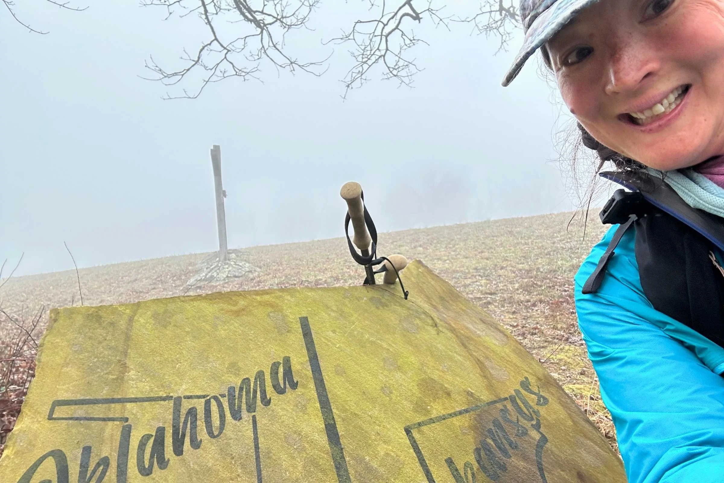 Liz Thomas smiling at the Oklahoma state line marker on a foggy day during her Ouachita Trail thru-hike, wearing a blue jacket and backpack