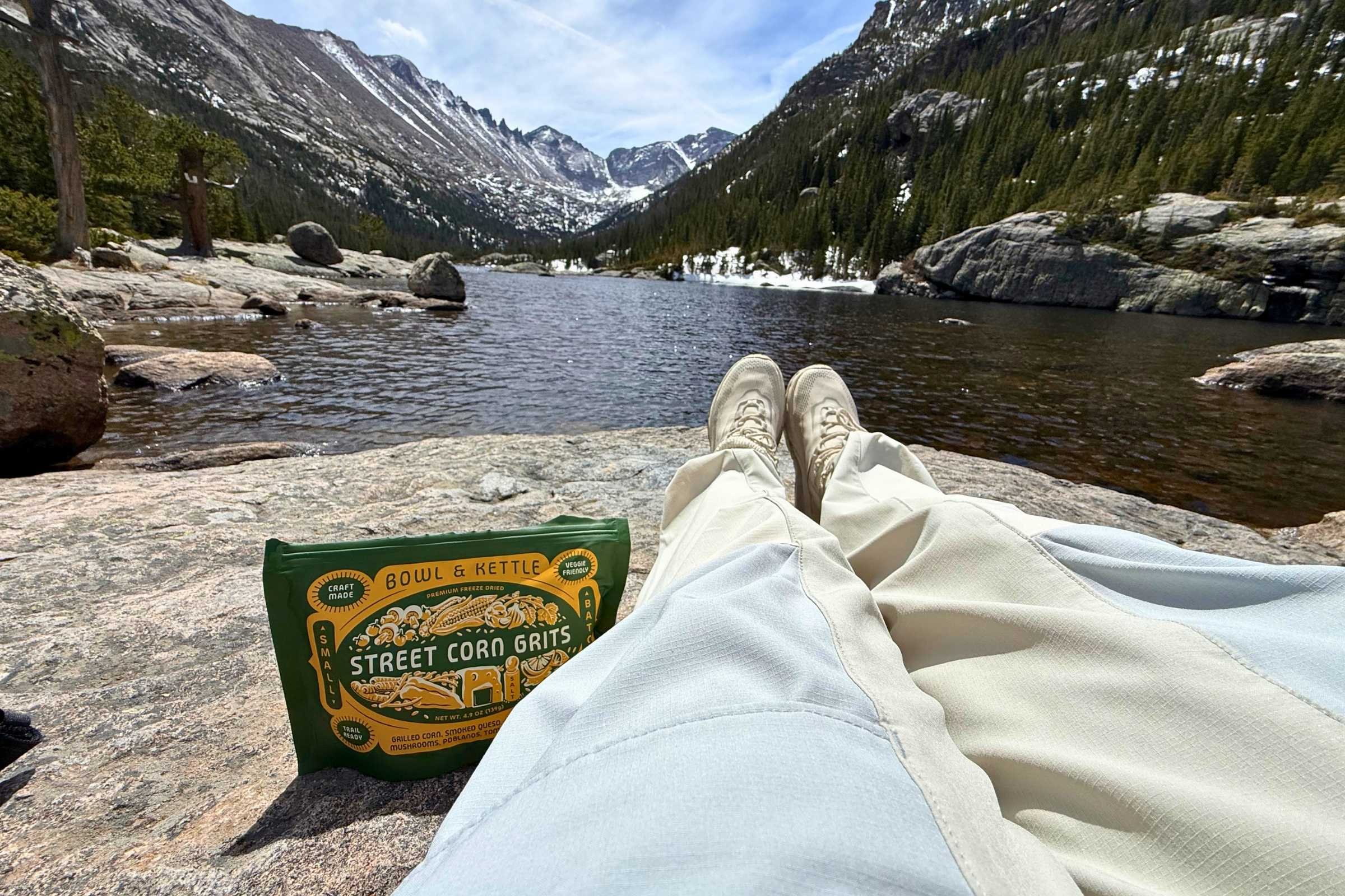 View of Bowl & Kettle Mexican Street Corn Grits freeze-dried meal next to a mountain lake with a person relaxing on the shore.