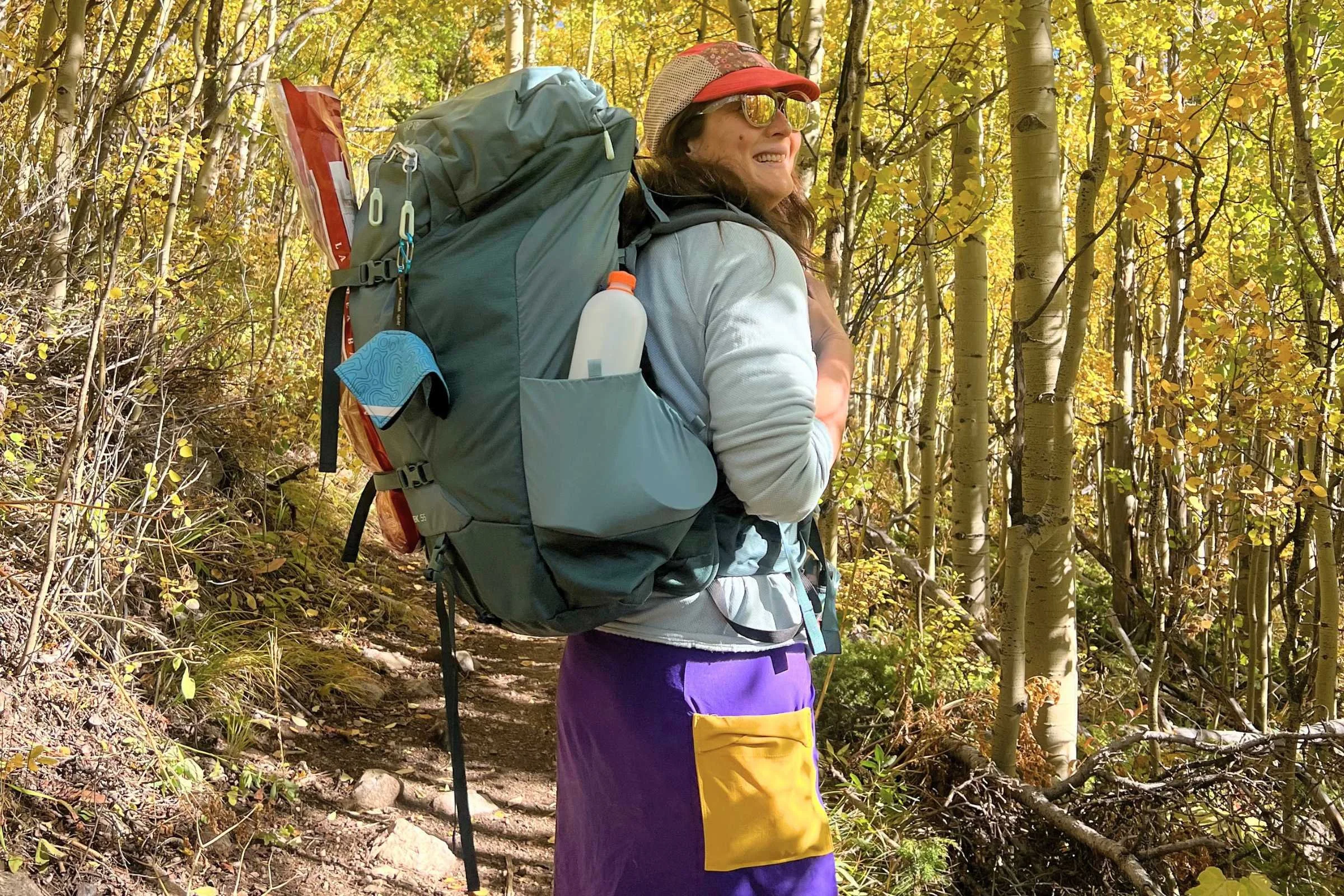 Woman wearing the Osprey Fairview Trek 55 Travel Pack while hiking through woods in a purple skirt.
