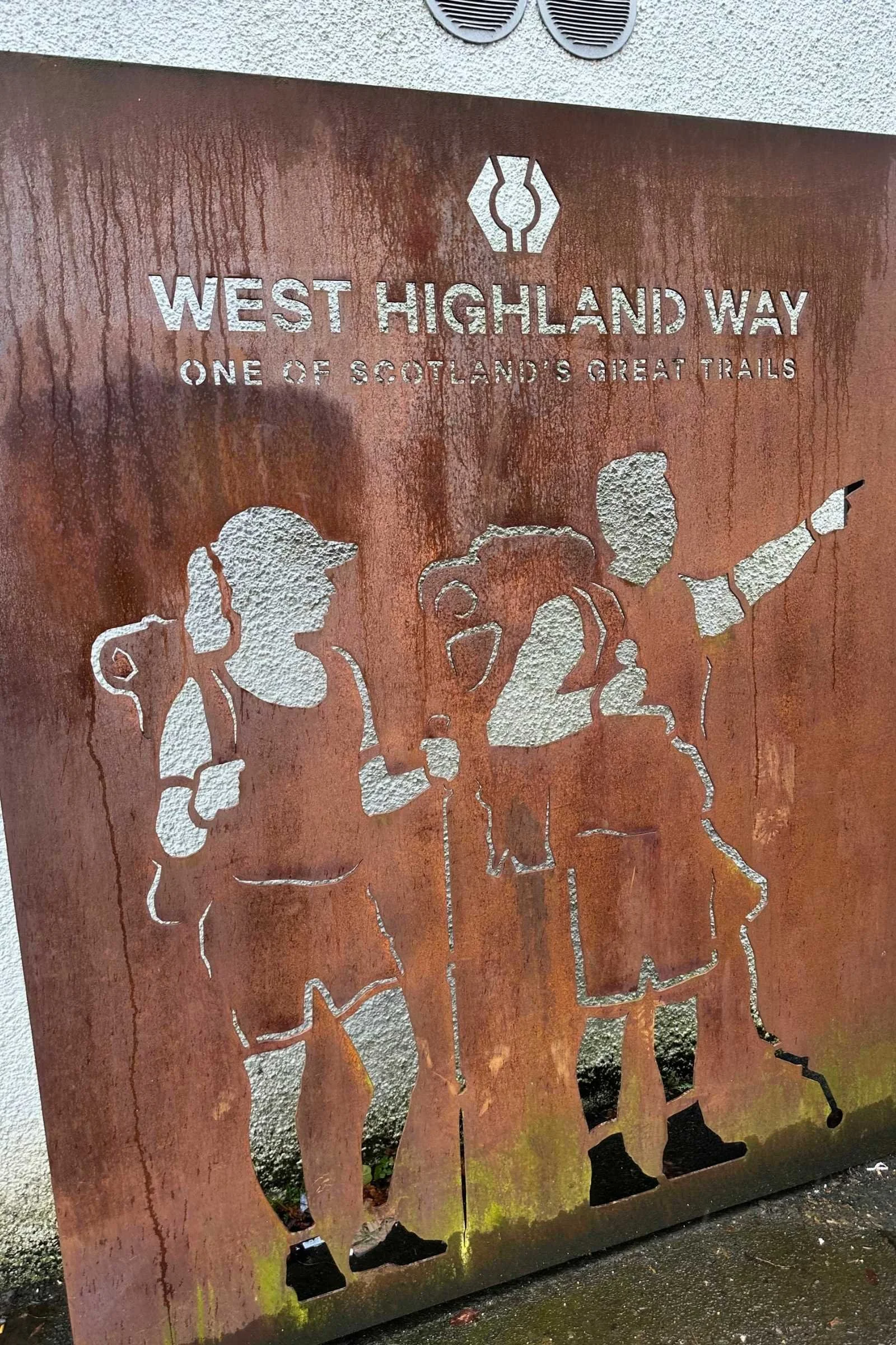 A large metal sign with an image of two backpackers looking ahead marks the start point of the West Highland Way.