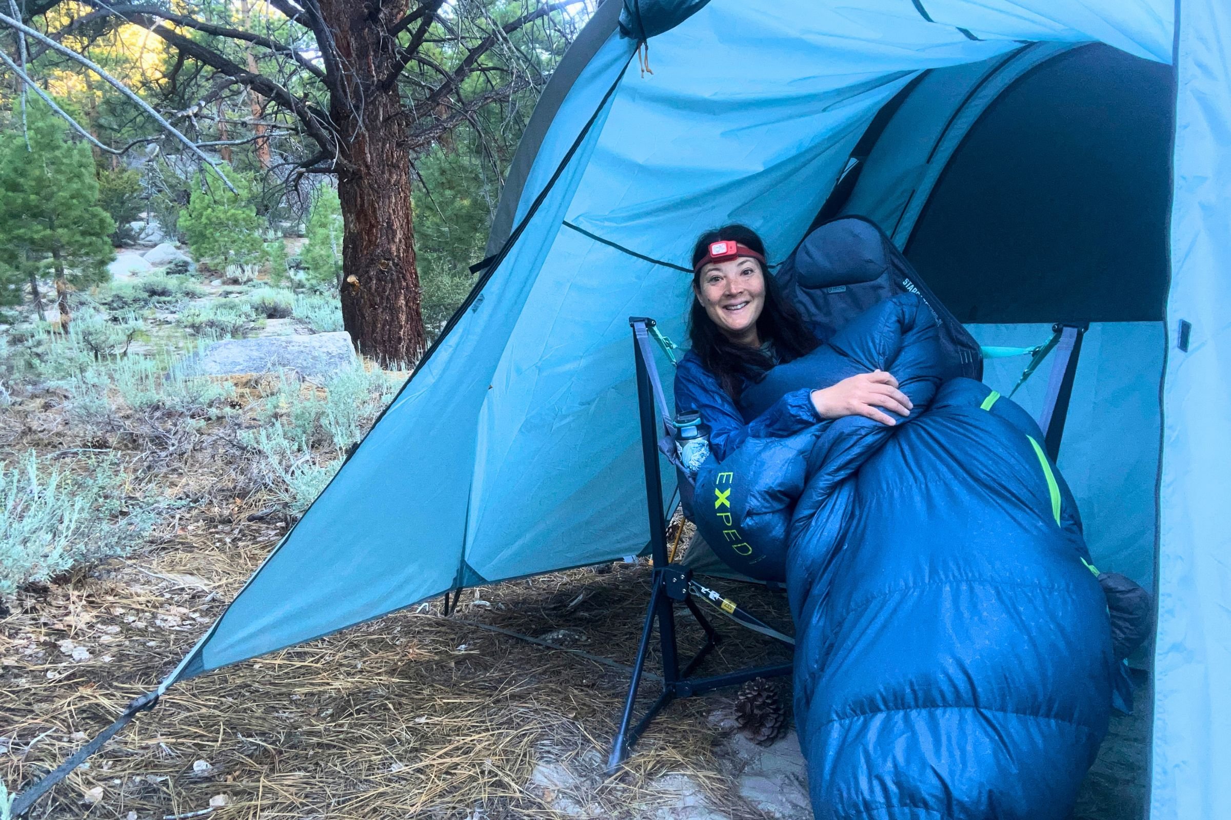 A woman sits inside an Exped Terra Sleeping Bag on a camping chair inside a tent in a wooded setting.
