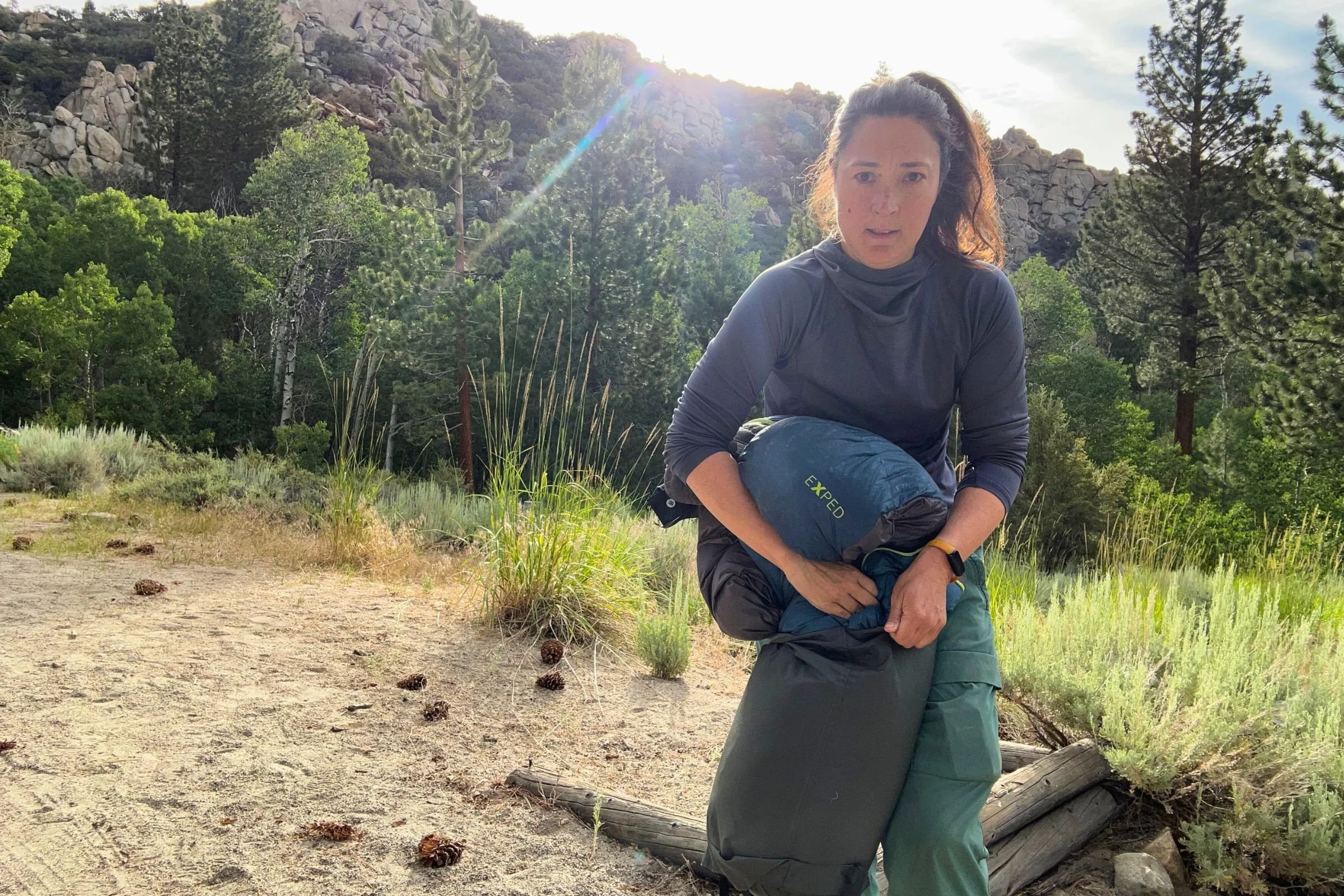A woman stuffing the Exped Terra sleeping bag into a stuff sack with trees in the background.