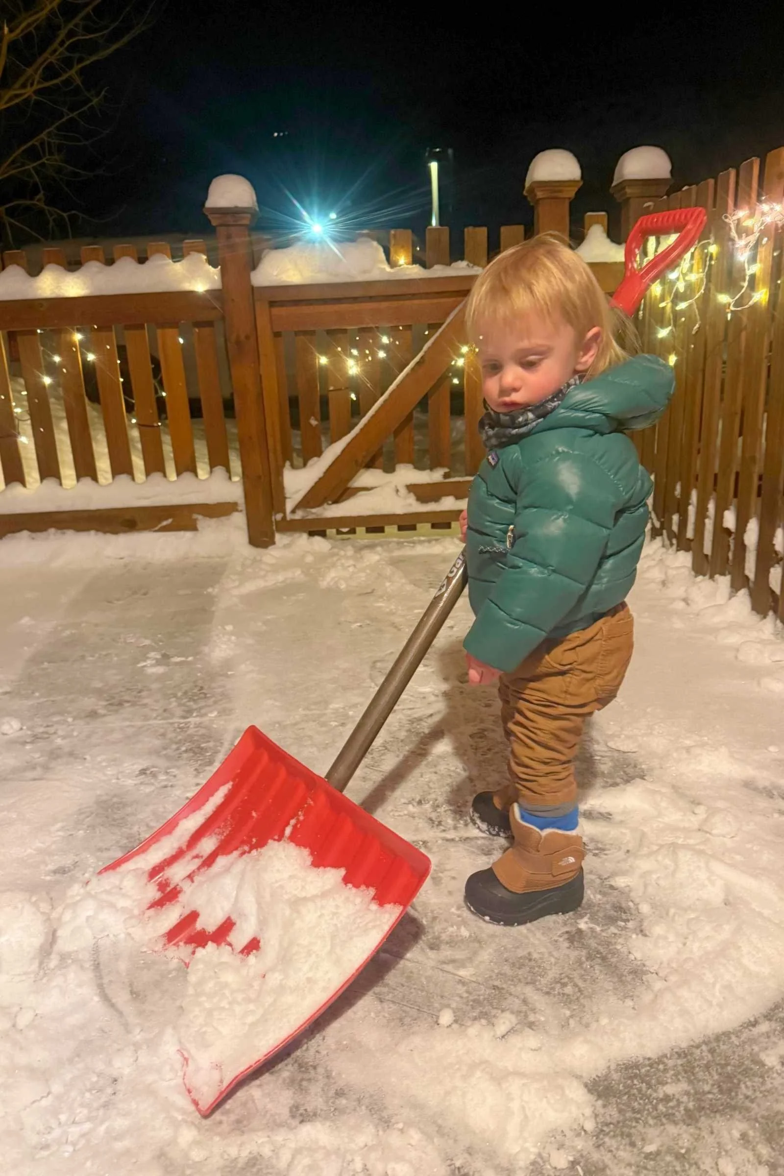 Cute toddler shoveling snow at night wearing The North Face Alpenglow II winter boots