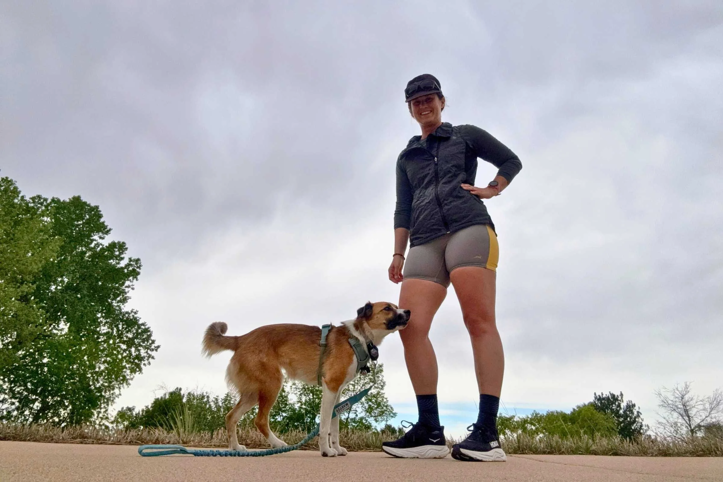 A person in Tracksmith Twilight Short Tights stands next to a dog on a path with trees and a cloudy sky in the background.