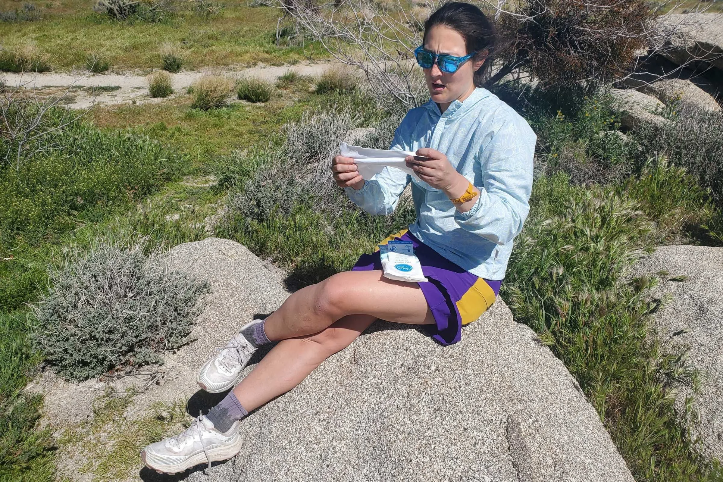 A woman sits on a rock in front of bushes and cactus. She holds a Cleanlife Bathing Wipe and has the package on her lap.