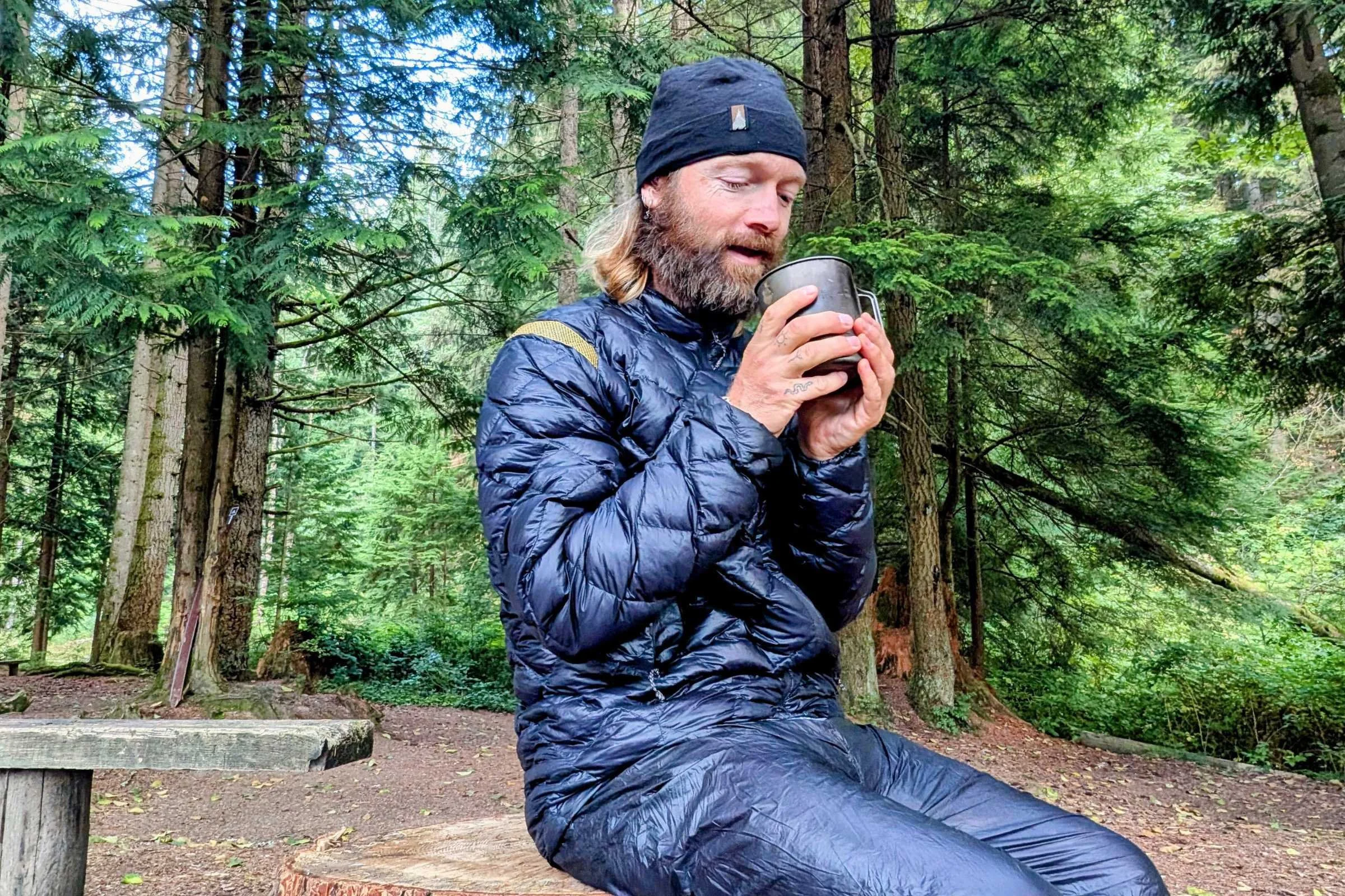 Camper sitting in forest wearing La Sportiva Lumina down jacket and holding mug