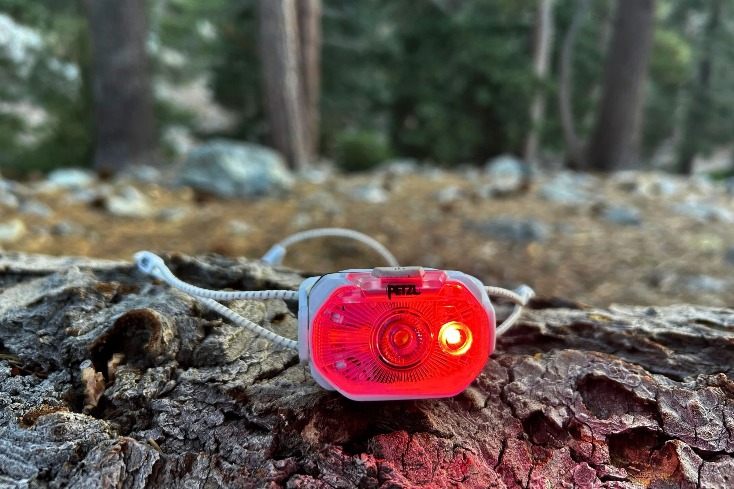 A closeup of the Petzl Swift LT headlamp in red light mode. It's sitting on a log, with trees and rocks in the background.