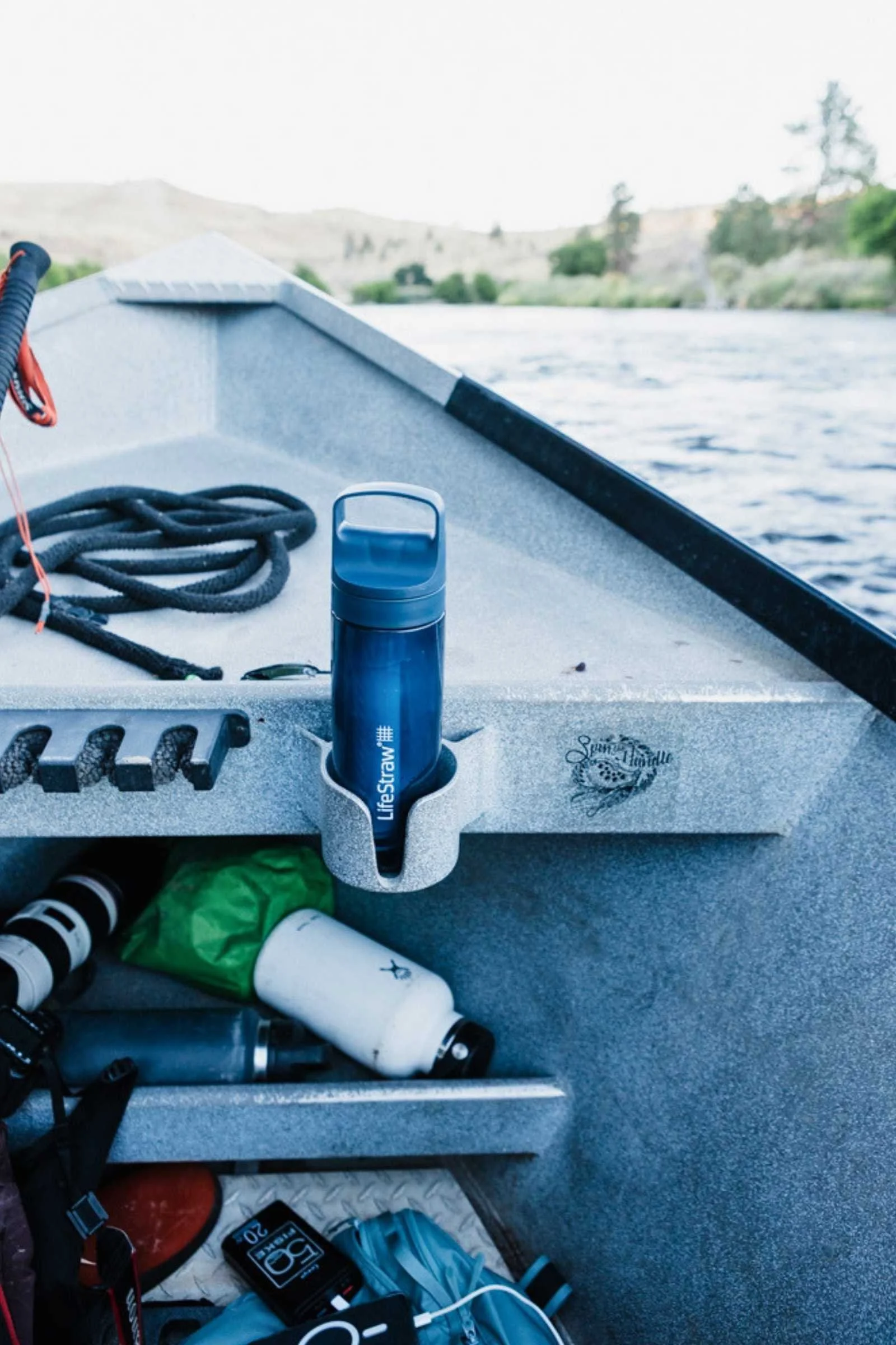 A blue LifeStraw bottle sits upright in a built-in holder on the side of a small aluminum boat. Coiled rope and other gear are stored nearby, with a calm lake and pale, tree-covered hills visible in the background.