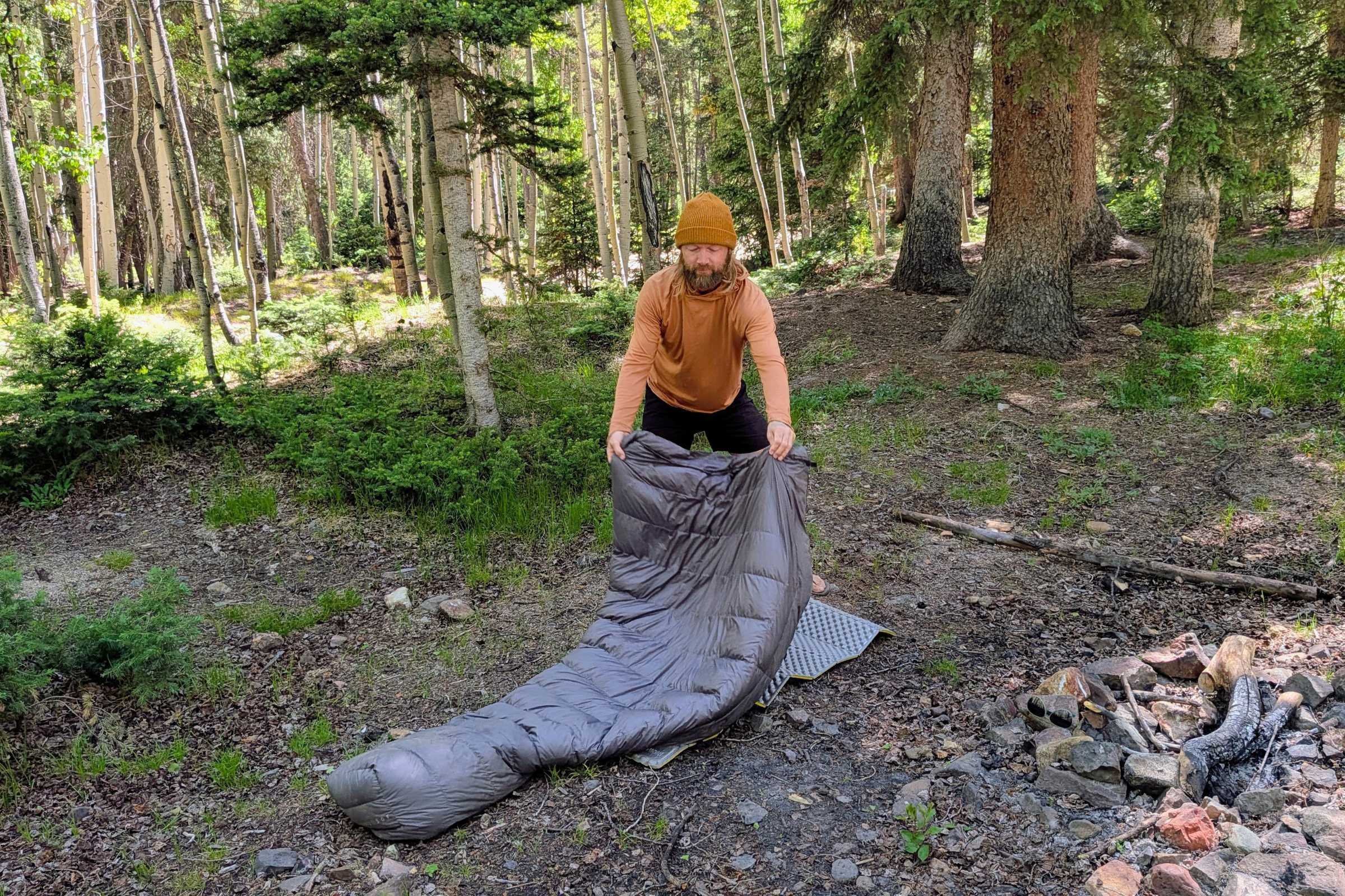 The author unfolds the Outdoor Vitals StormLoft 15 Down TopQuilt in a forest next to a fire ring