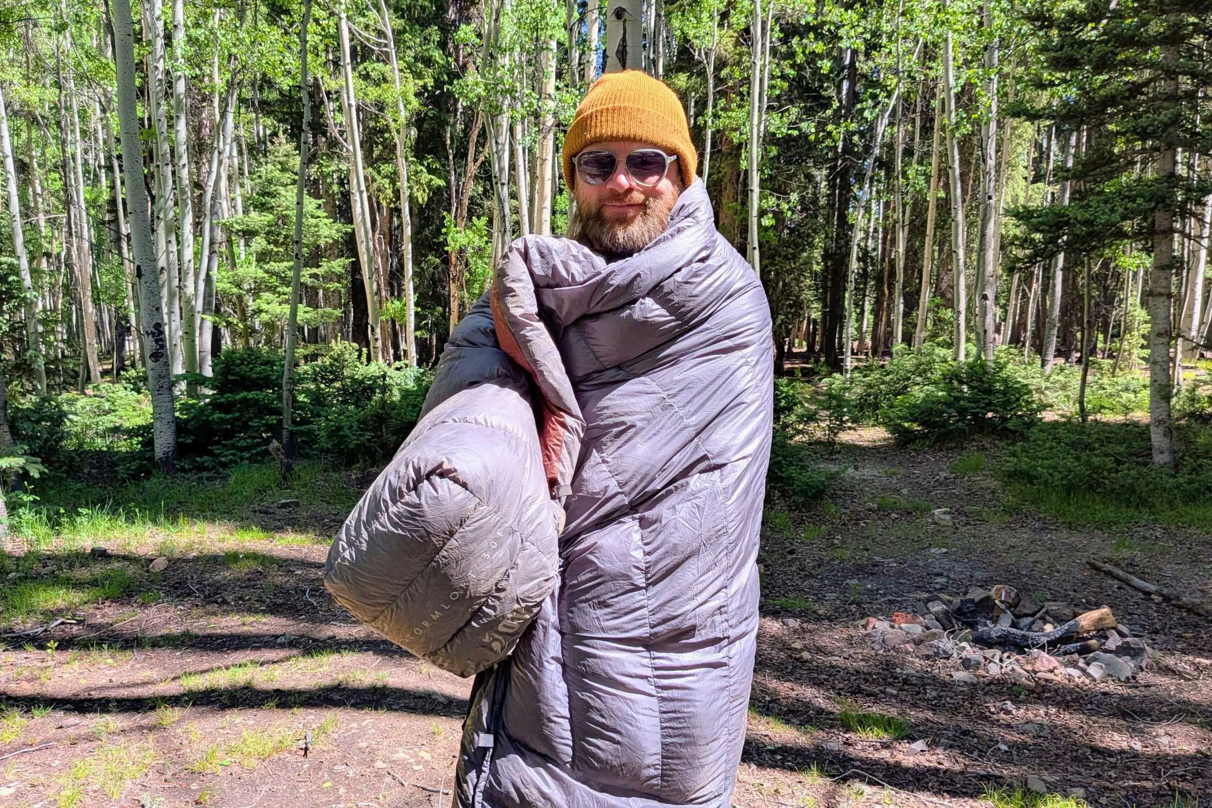 Author Sam Schild wearing a hat and sunglasses while wrapped in Outdoor Vitals StormLoft 15 Down TopQuilt in a forest