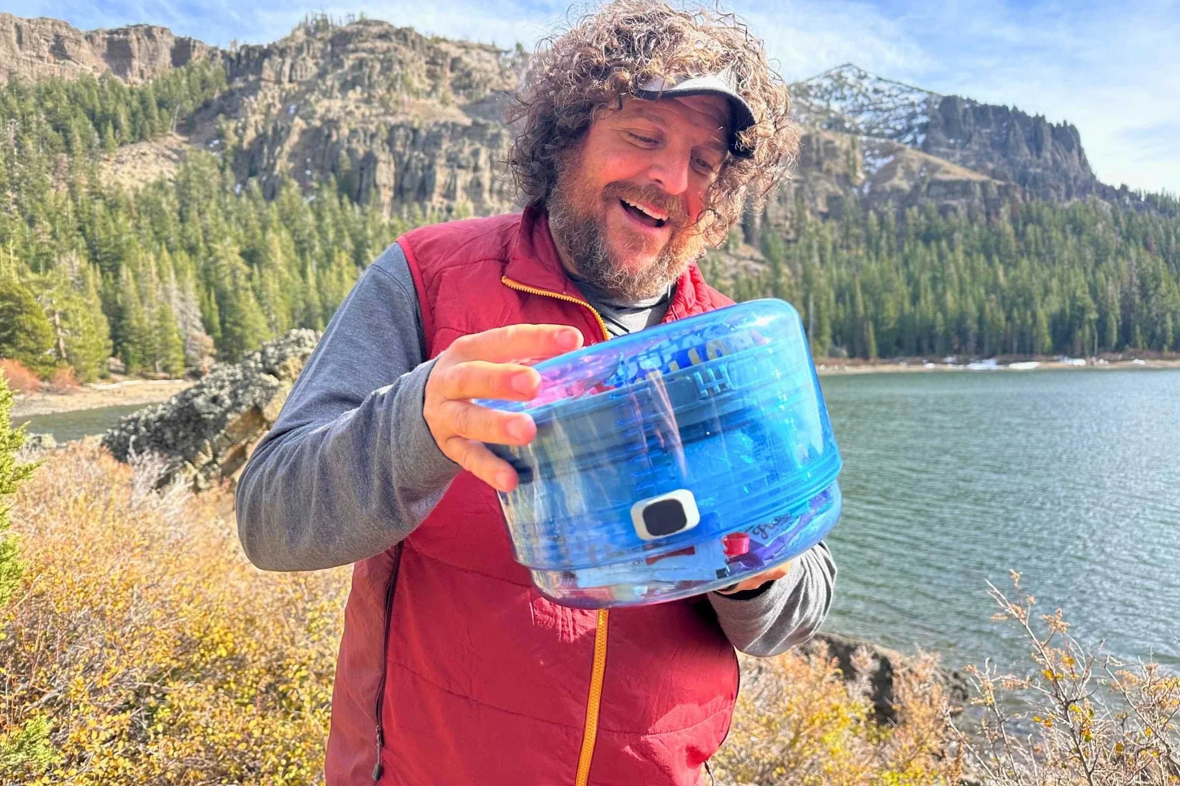 Man holding the Bearvault BV One bear canister by a body of water with the mountains in the backgroundPhoto courtesy Sara Kruglinski.