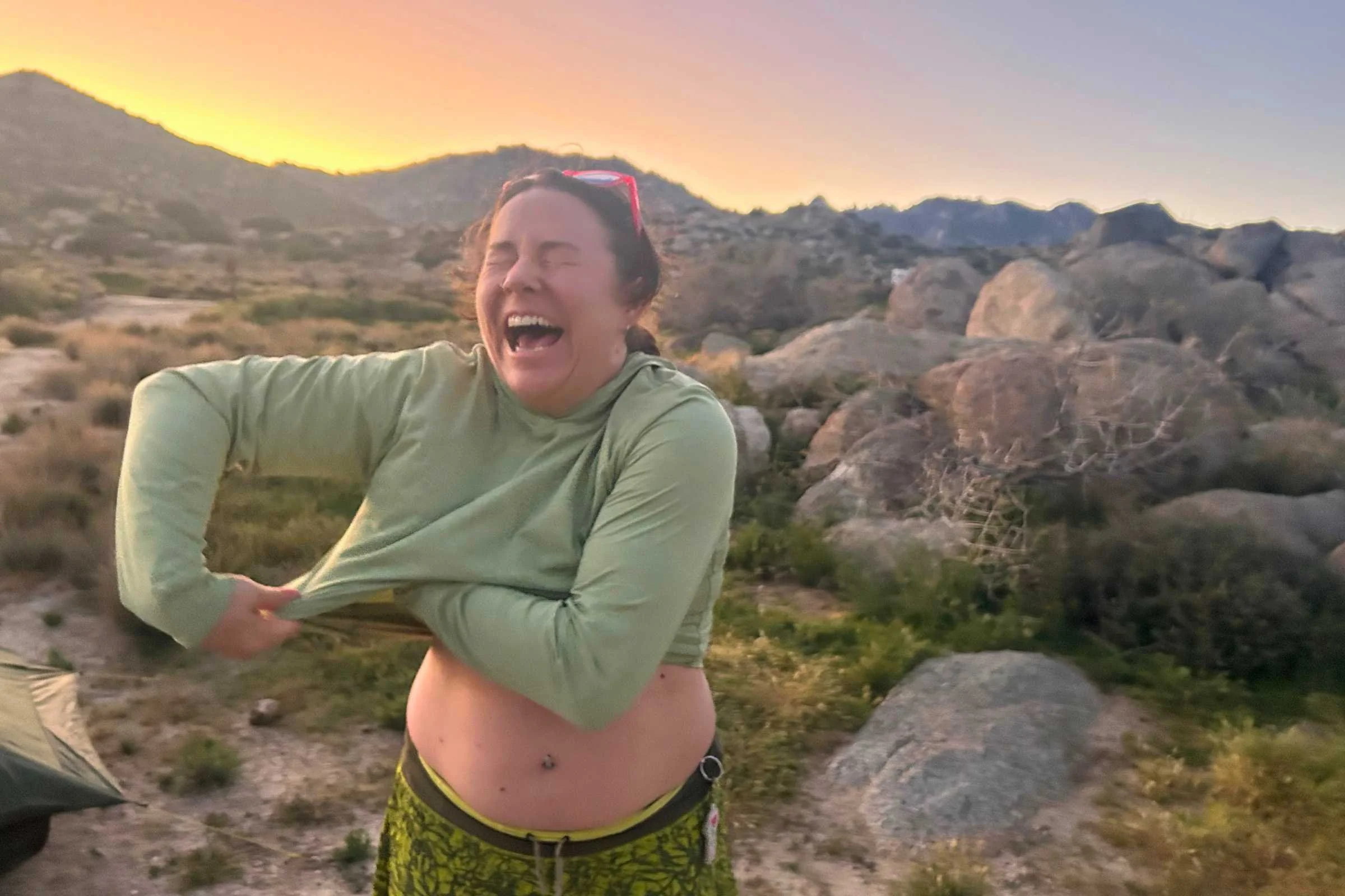 A laughing woman cleans her underarms with a cleansing wipe in front of a sunset landscape with hills and rocks.