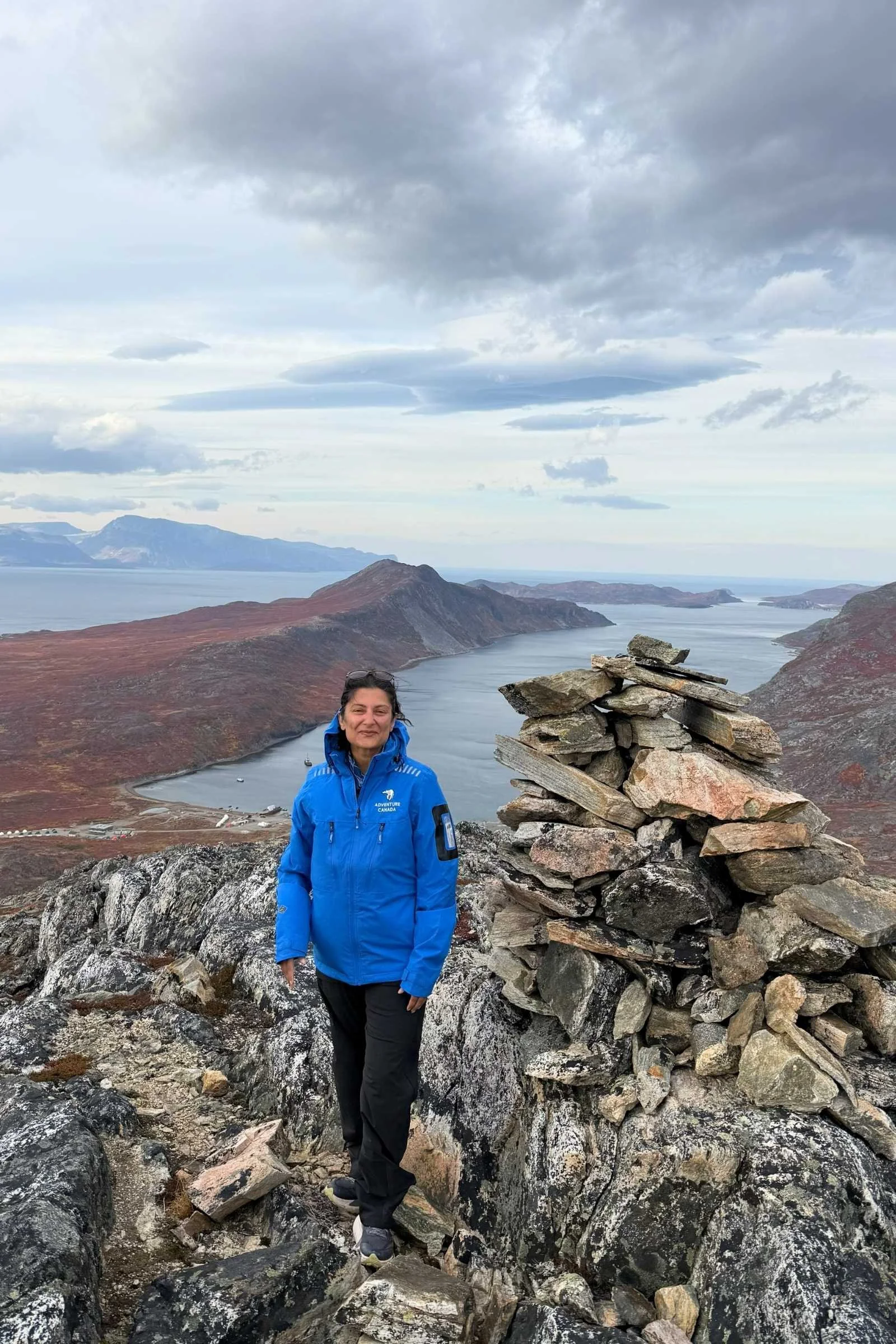the author in a blue raincoat on a mountain with a lake and gray clouds in the background