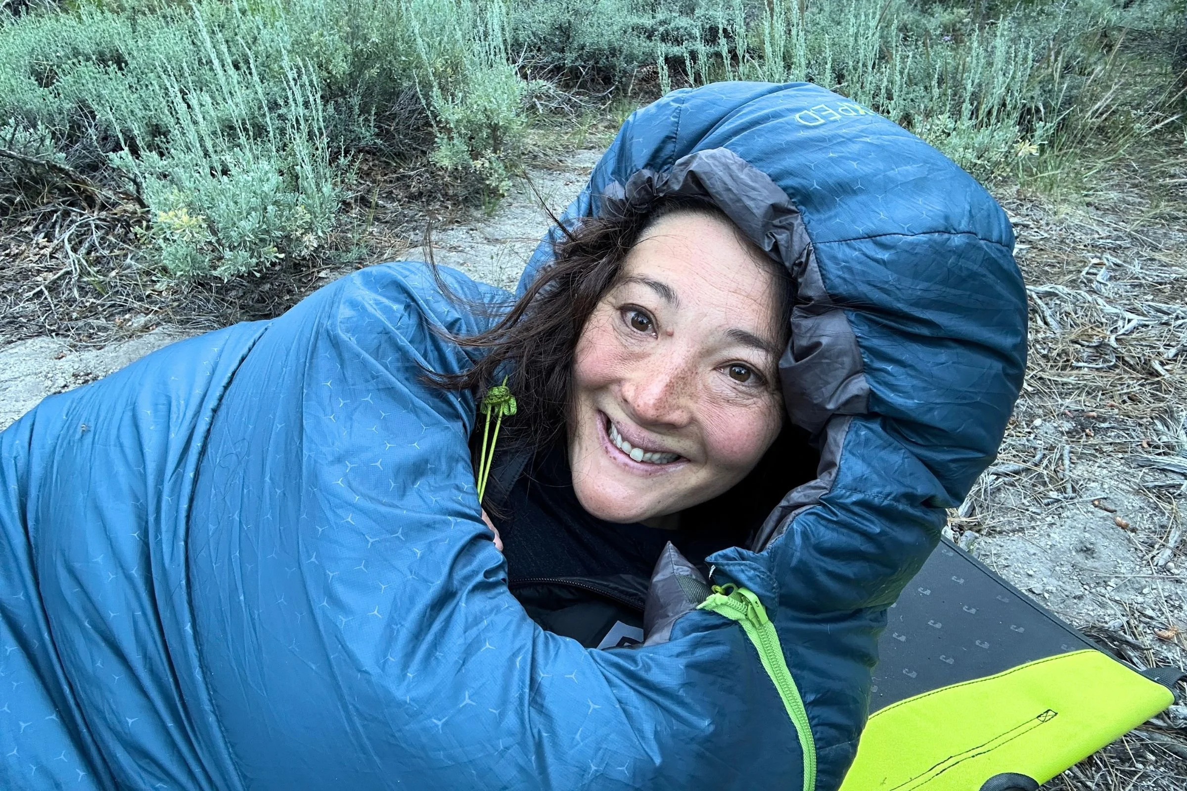 A woman lies on the ground inside an Exped Terra Sleeping Bag outdoors.
