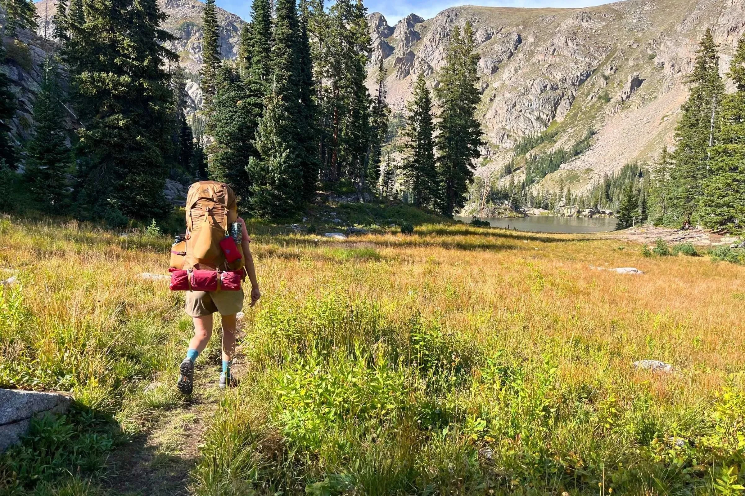 A person wearing a backpacking backpack hiking on a trail through a meadow to the edge of an alpine lake