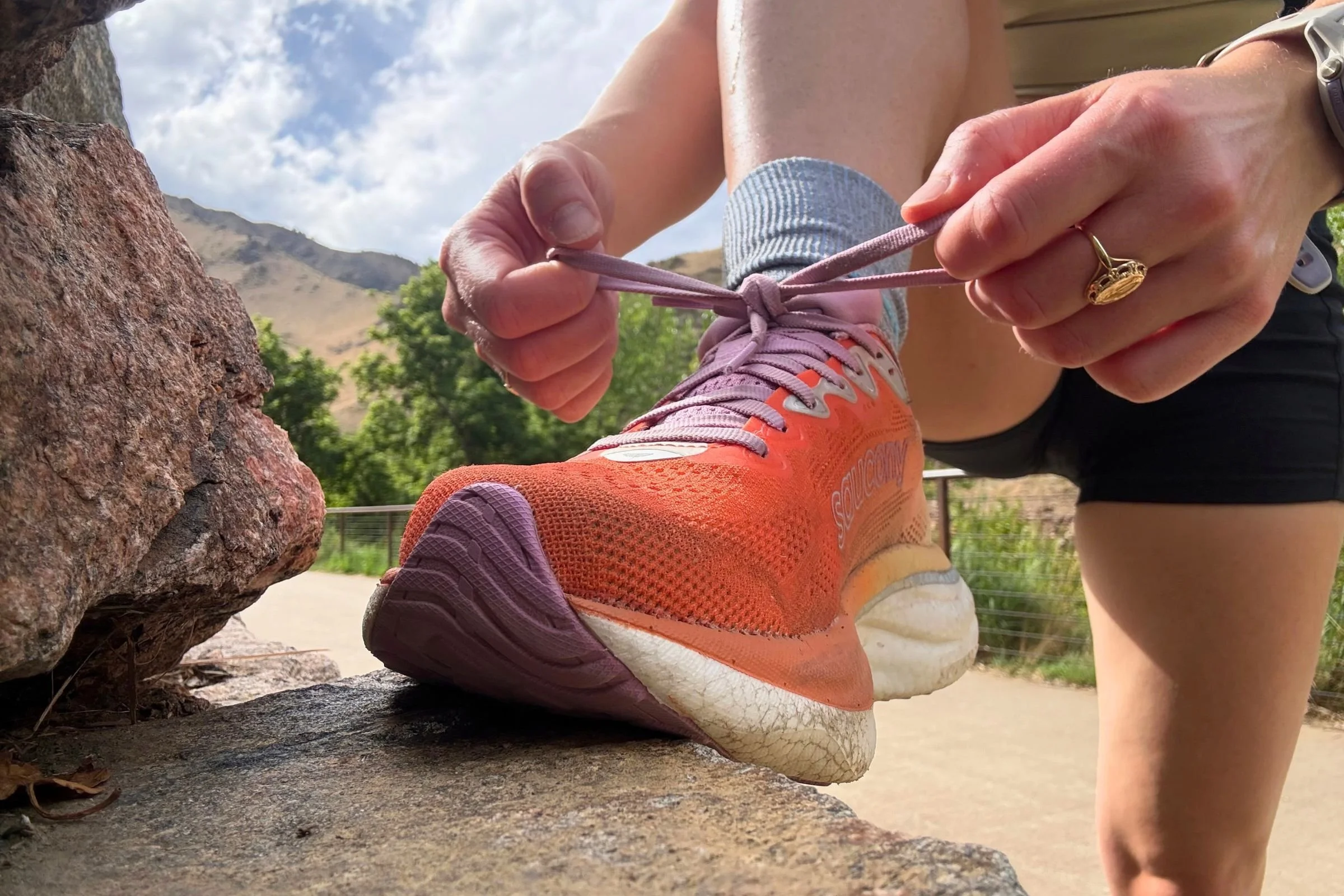 A woman ties her Saucony Ride 19 Road Running Shoes while standing next to a rock.