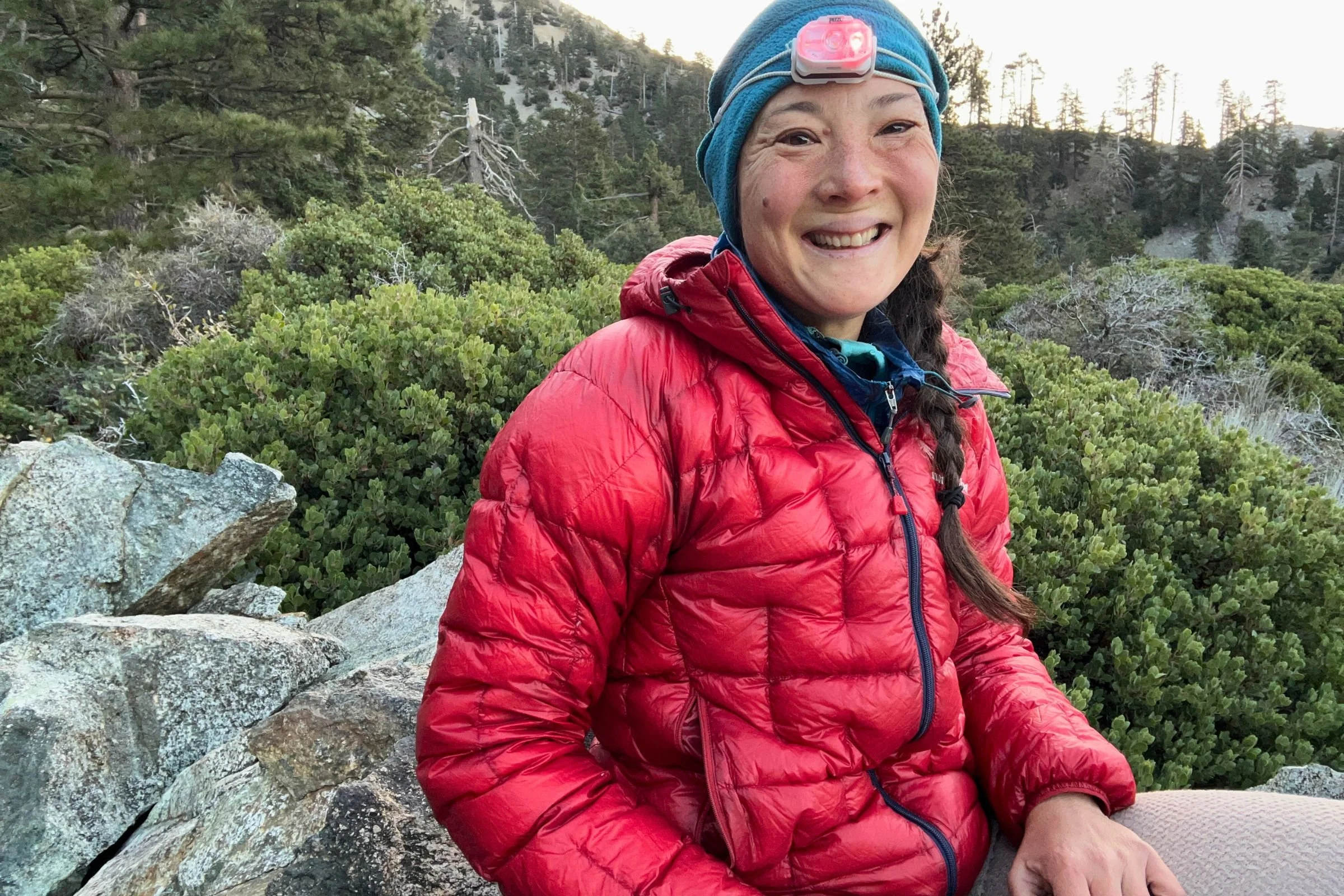 The author, Liz Thomas, wearing a red down jacket, wearing the Petzl Swift LT headlamp, with shrubs and rocks in the background.