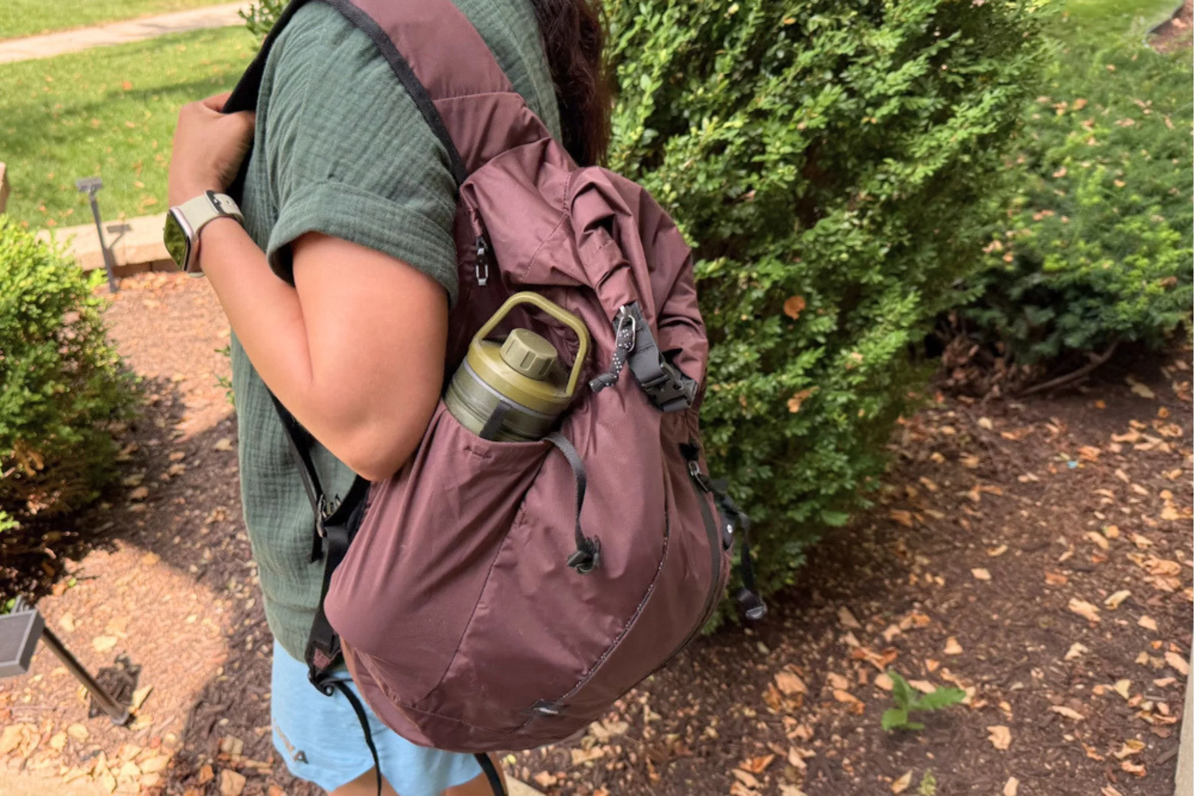 A person wearing a green shirt and a maroon backpack with Grayl UltraPress Filter and Purifier Bottle in the side pocket.