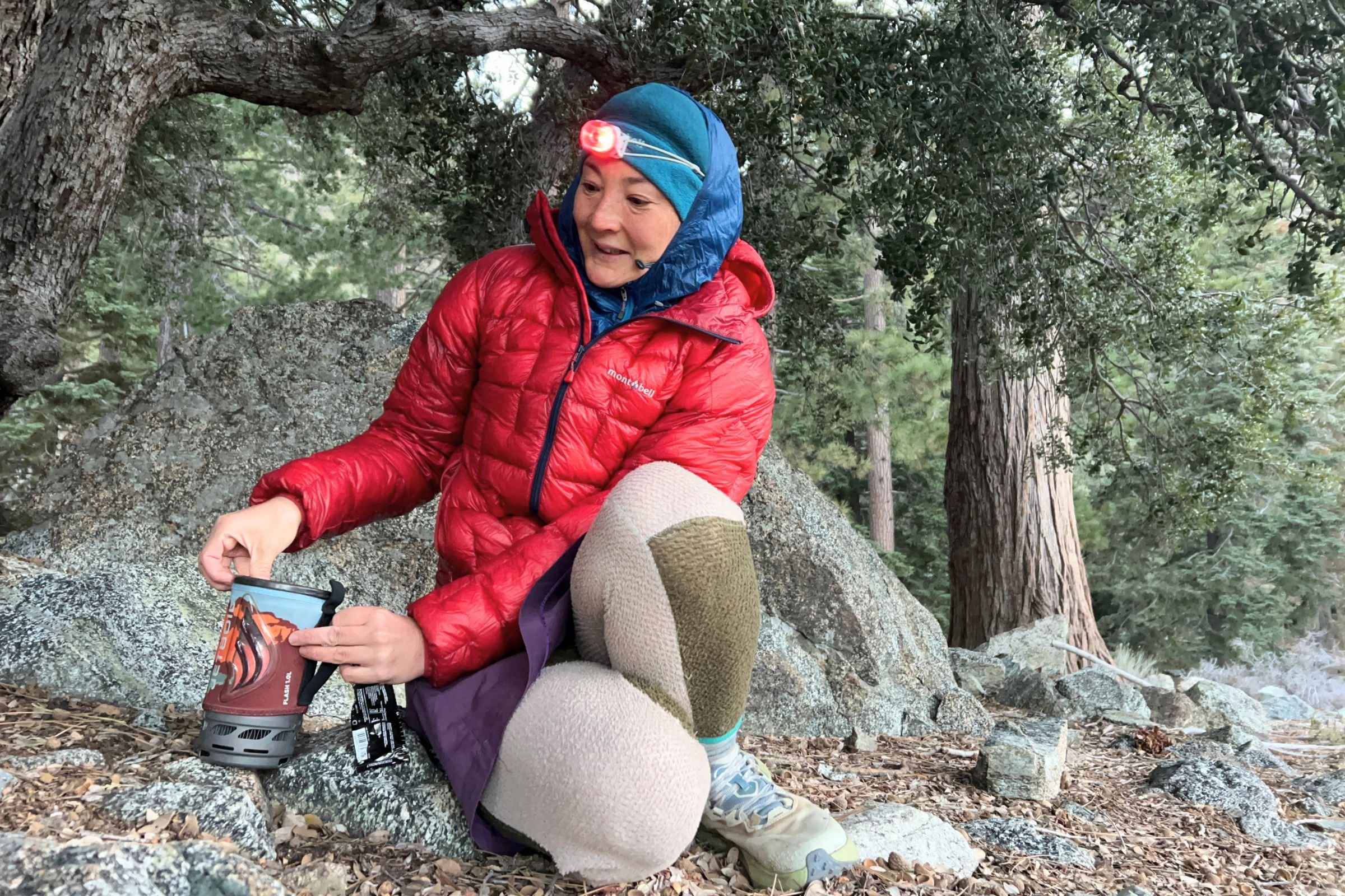 The author wearing a red down jacket and fleec pants on a cold, early morning backpacking trip. She is wearing the Petzl Swift LT on top of a blue fleece cap.