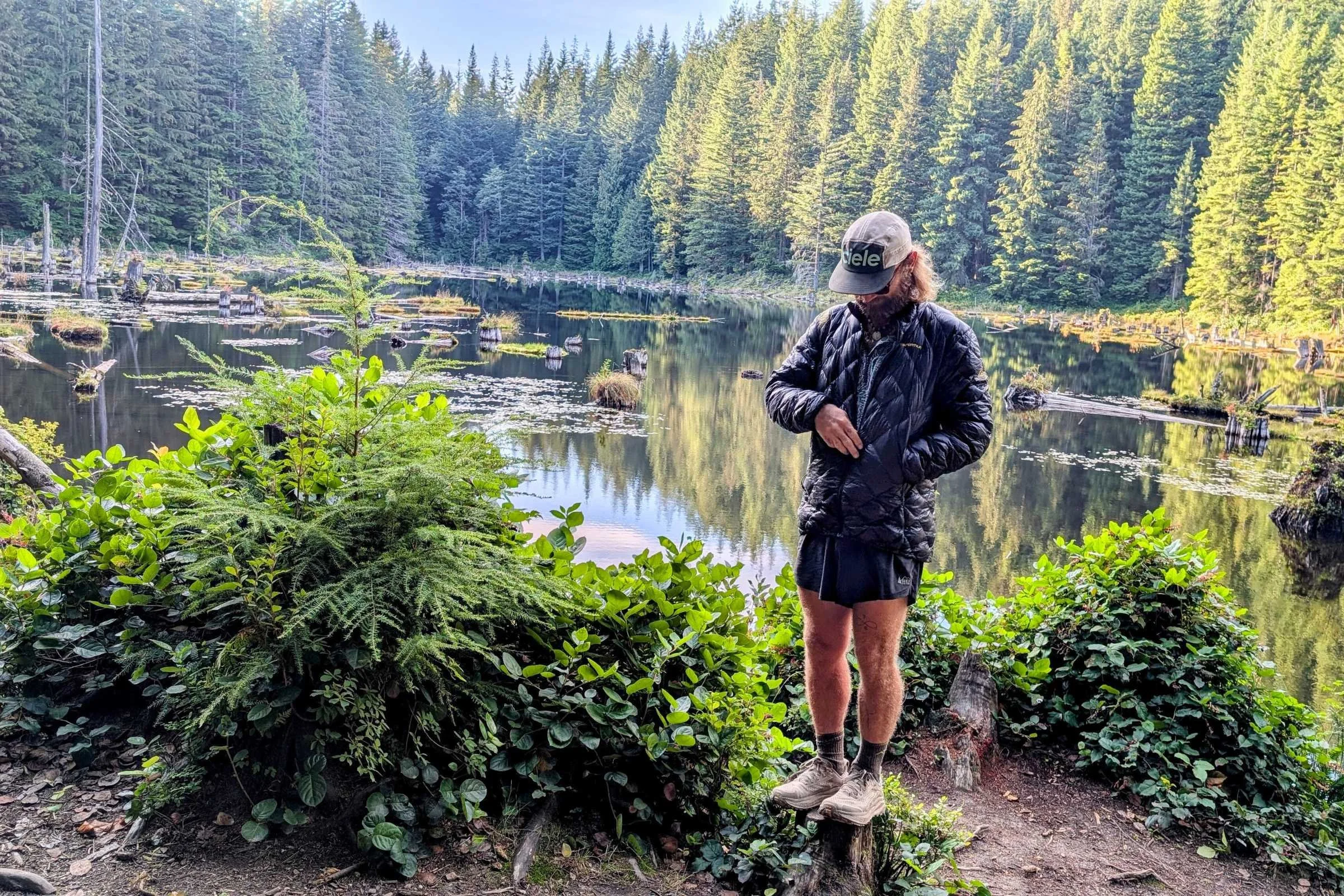 Man standing by forest lake wearing La Sportiva Lumina down jacket while adjusting zipper