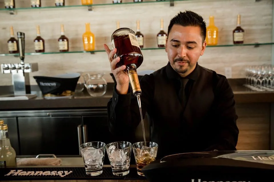 a young owner of Mobile Mixologist expertly pouring Hennessy into glasses lined up on a bar at a brand activation in a black suit