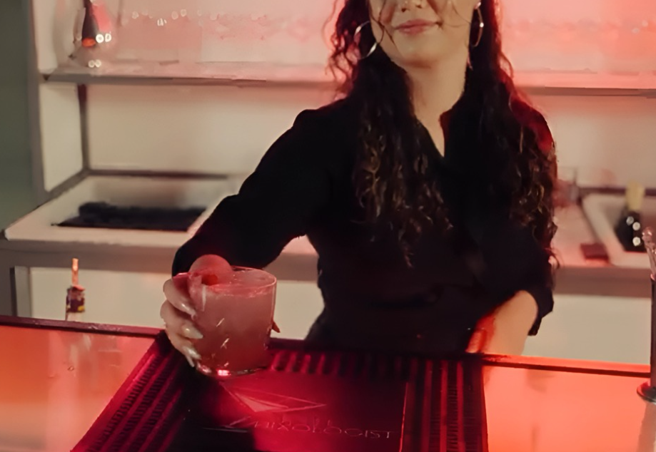A woman with curly hair and hoop earrings, wearing a black top, holds a cocktail glass with a pink drink at a bar with pink lighting.