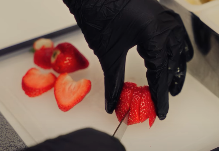 A person wearing black gloves slicing strawberries on a white cutting board.