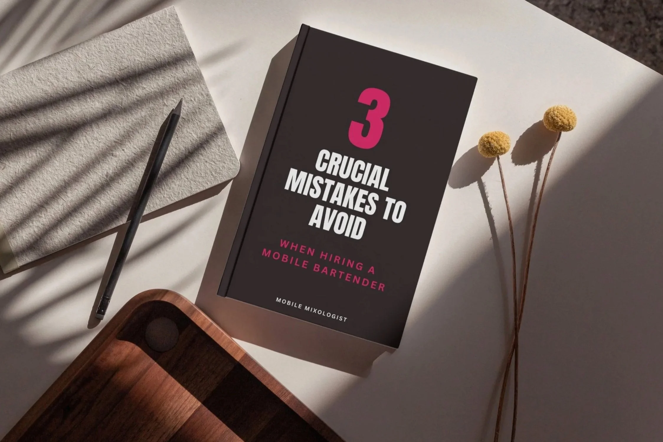 Mobile Mixologist Free E book on 3 Crucial mistakes to avoid when hiring a bartender, on a desk with a pen, paper and flowers, for an elegant bar at your next party