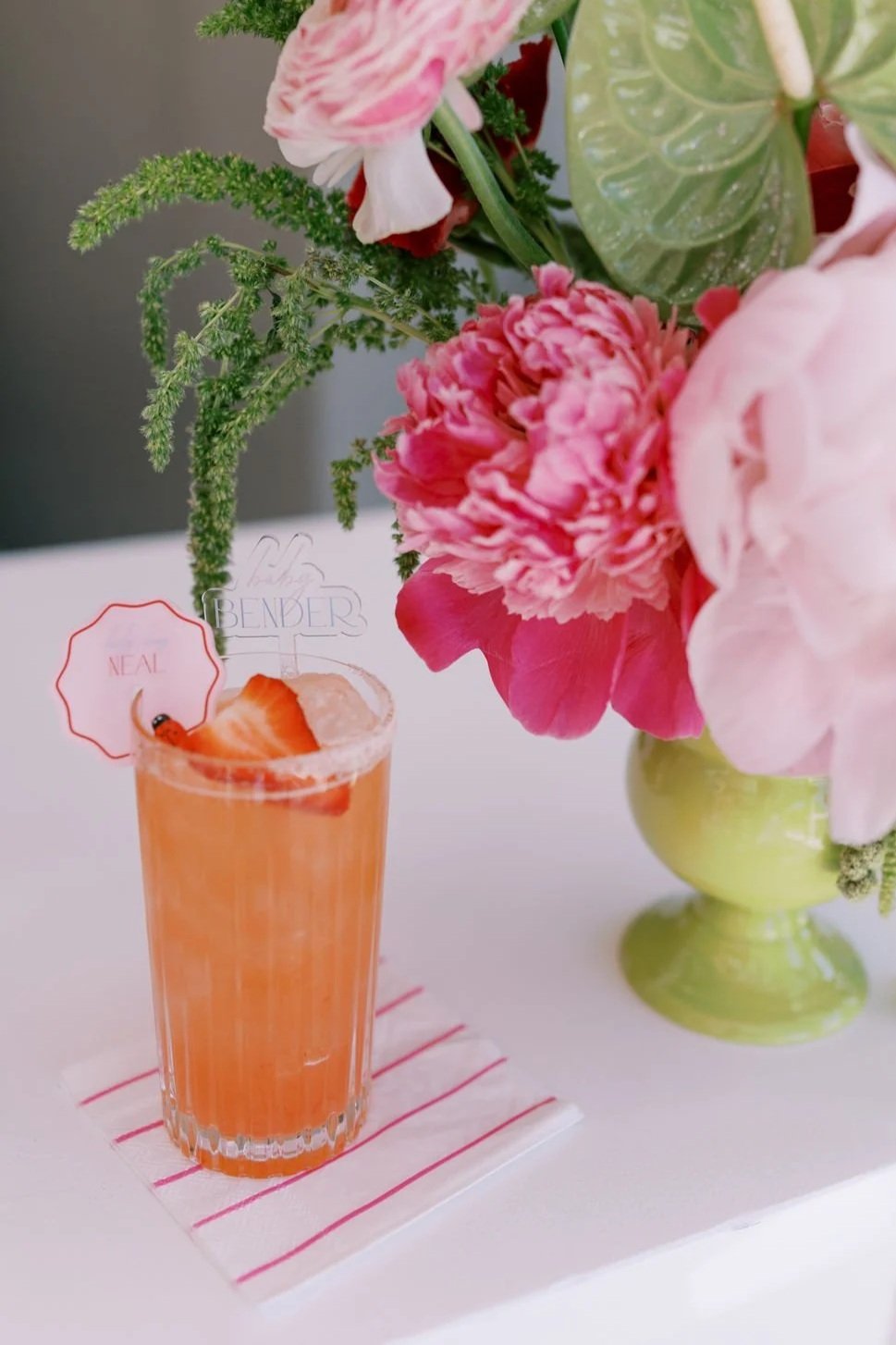 A pink cocktail with a strawberry slice and a small sign reading 'Neal' in front of a floral arrangement with pink and white flowers in a green vase.