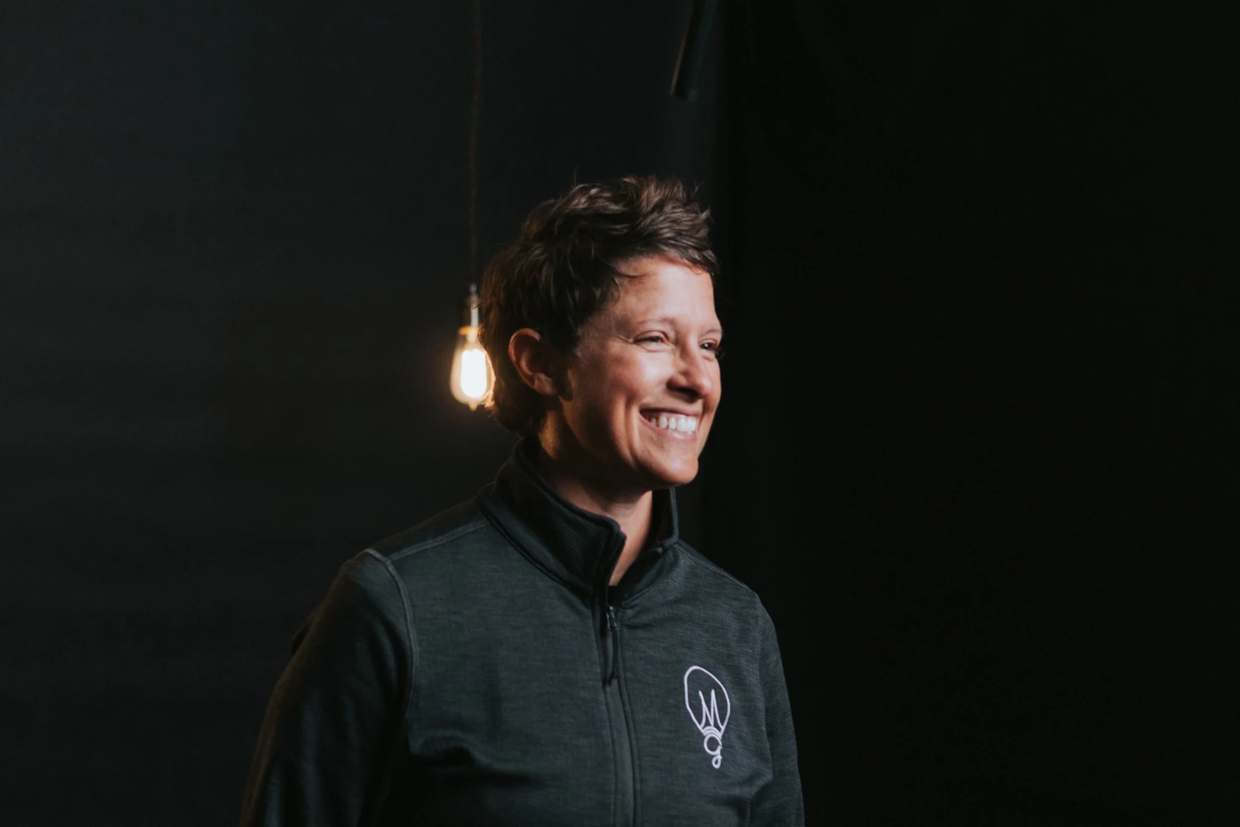 Smiling woman with short dark hair wearing a dark jacket with a logo, standing against a dark background with a visible light bulb.