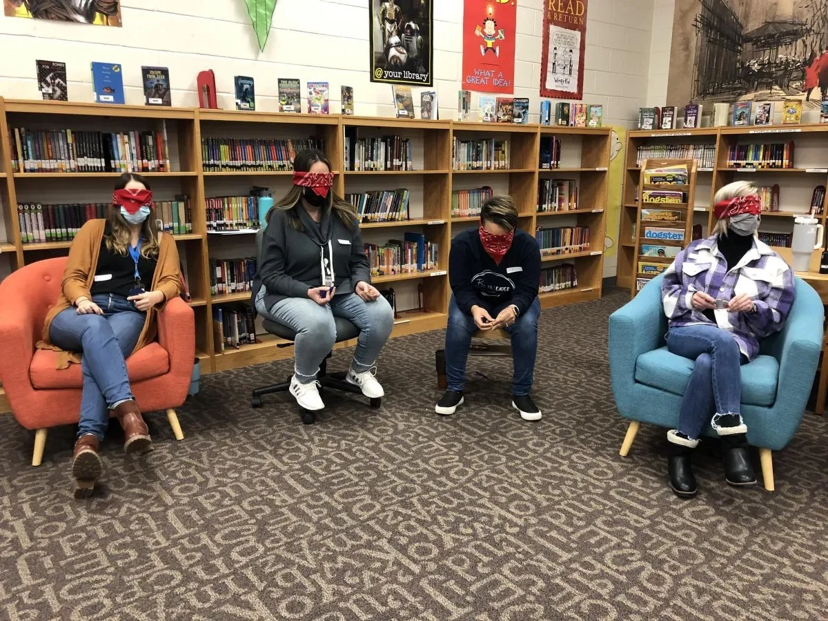 Four people sitting in a library, wearing face masks and red bandanas as blindfolds. The two on the left are seated in orange and black armchairs, and a person in the middle is seated on a black chair, with a purple armchair on the right. Behind them are bookshelves filled with books, and posters are hanging on the wall.