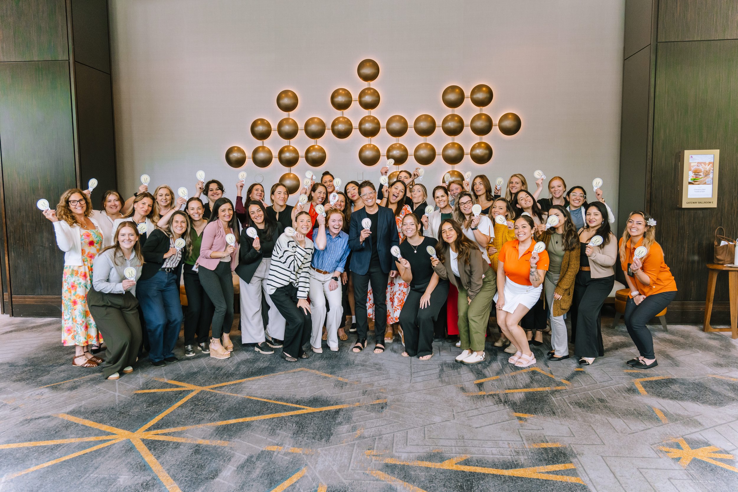 A large group of women and a man pose together at an indoor event, holding up signs with the 'M' logo, standing in front of a decorative wall with large spherical ornaments.