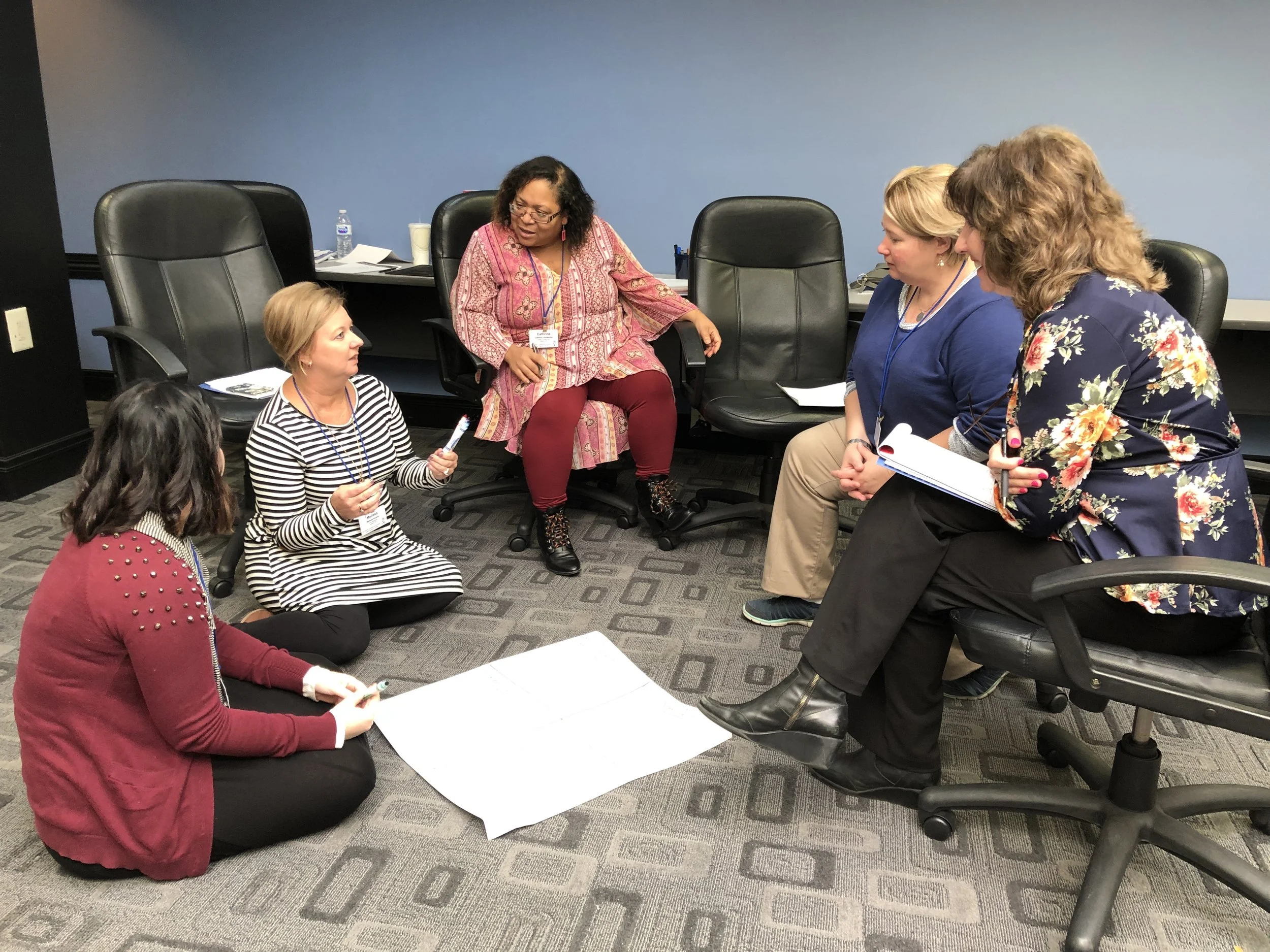 A group of women in a conference room having a discussion, some are seated on the floor while others are sitting on office chairs, with notebooks and papers in front of them.