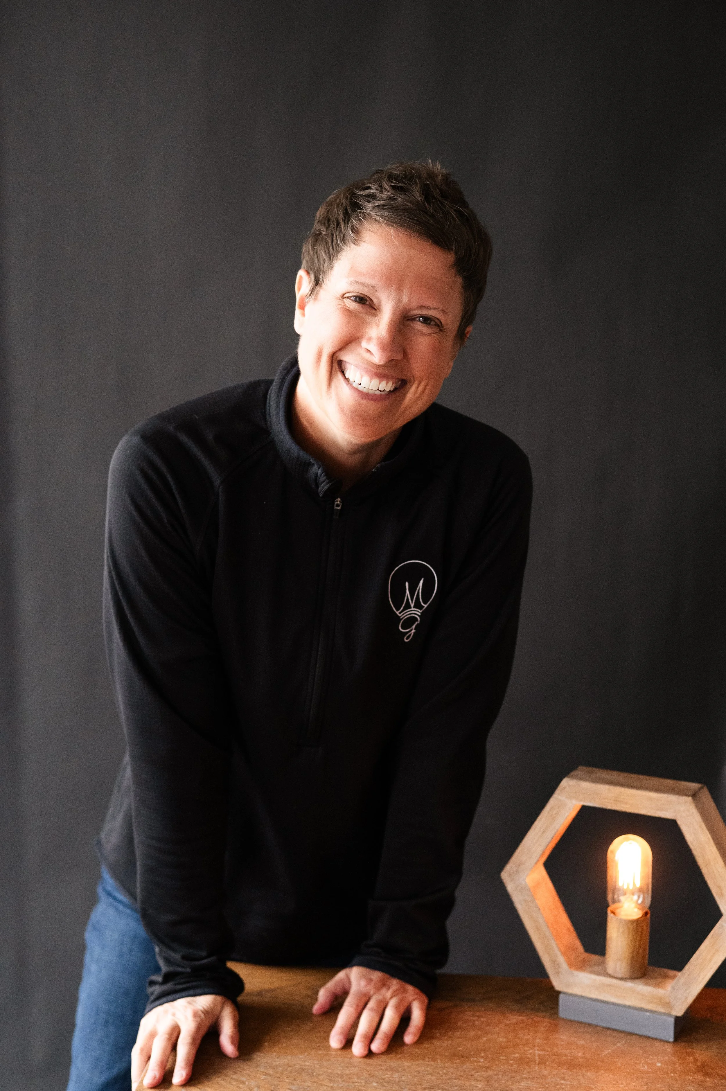 A smiling woman with short brown hair wearing a black zip-up jacket with a logo, standing next to a hexagonal wooden lamp with an exposed Edison bulb, against a dark gray background.
