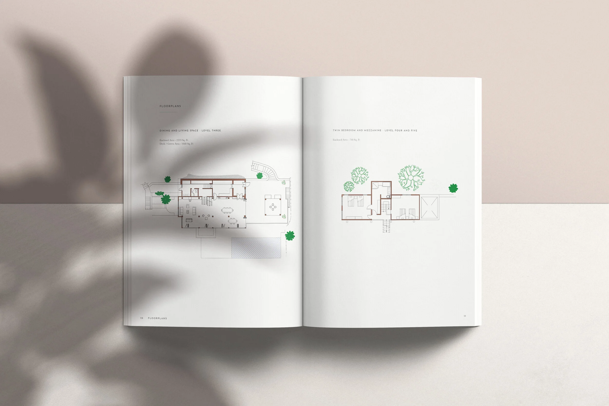Open book displaying floor plans of a house, with rooms and outdoor areas marked, and green tree illustrations, on light-colored surface with a shadow of a plant.