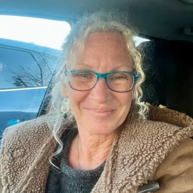 A smiling older woman with curly white hair and blue glasses, wearing a brown fleece jacket, sitting inside a vehicle