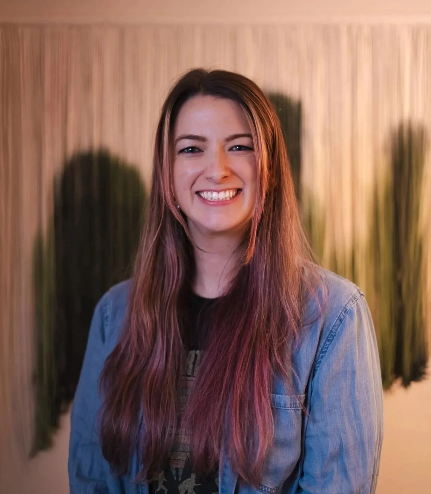 A young woman with long, pink-tinted hair, smiling with teeth showing, wearing a denim jacket, standing in front of a wooden wall and potted plants.