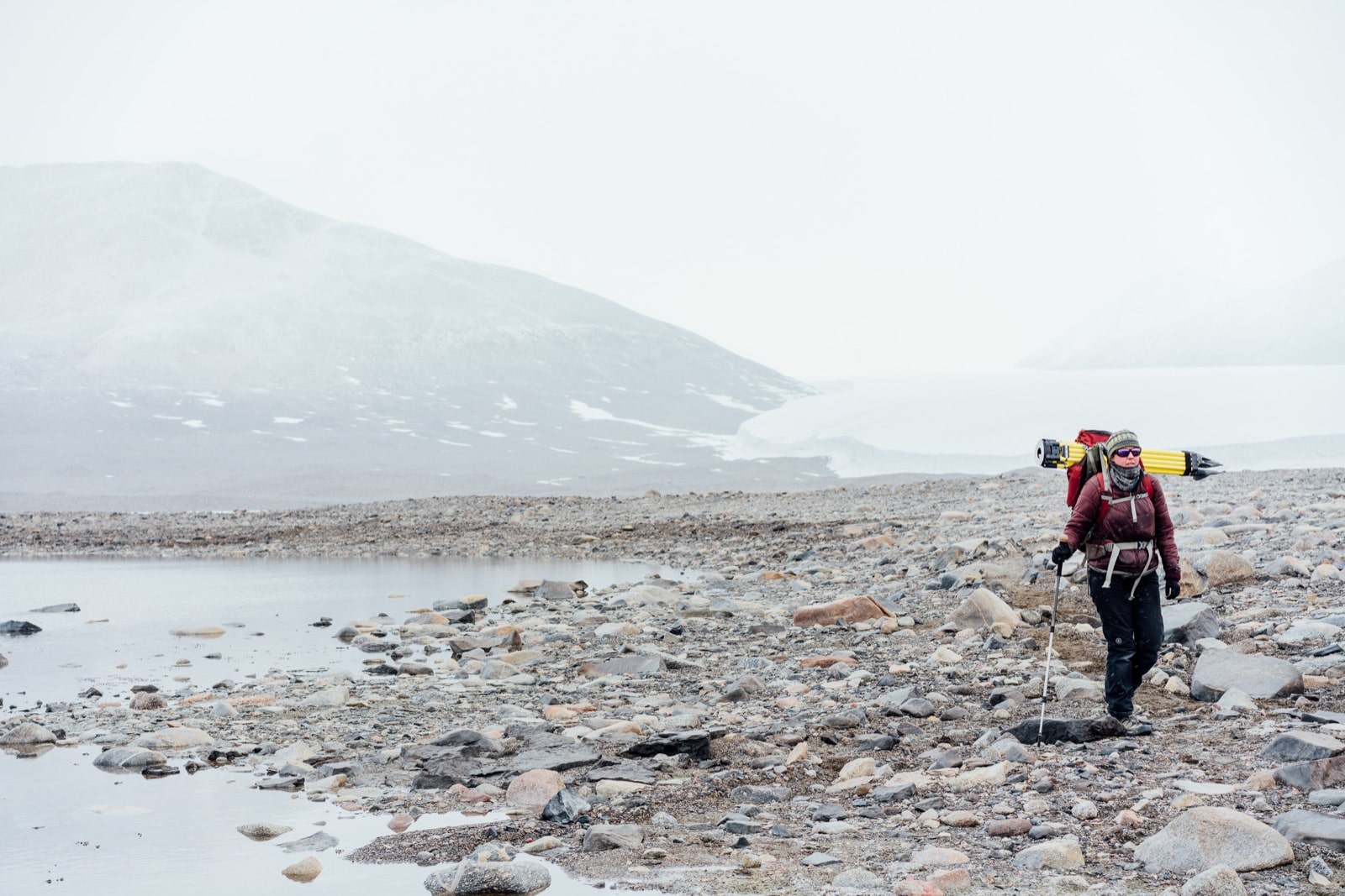  a woman carrying a lot of gear hikes in a rocky barren terrain. to the left is a small bank of water and behind is the edge of a glacier. 