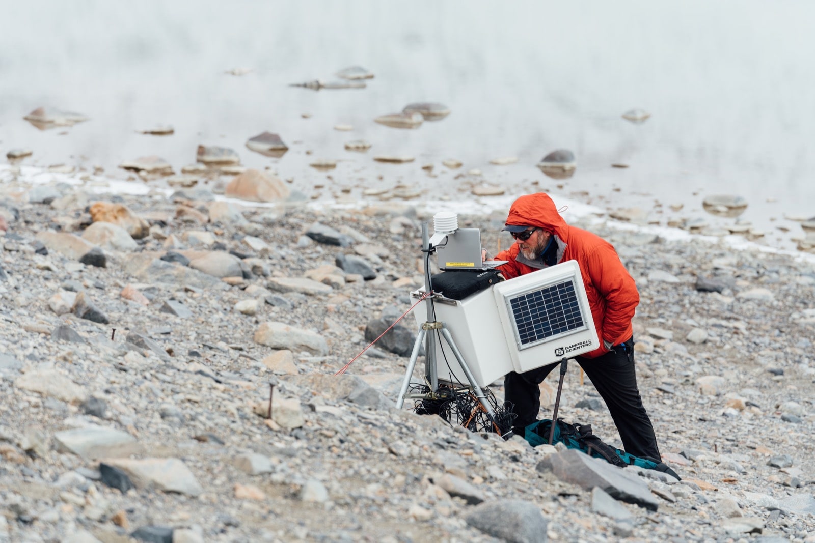  Mike Gooseff with his light red overcoat hood up over his head, working a box of lab equipment. He is standing in front of a bank of water on a rocky shore. 