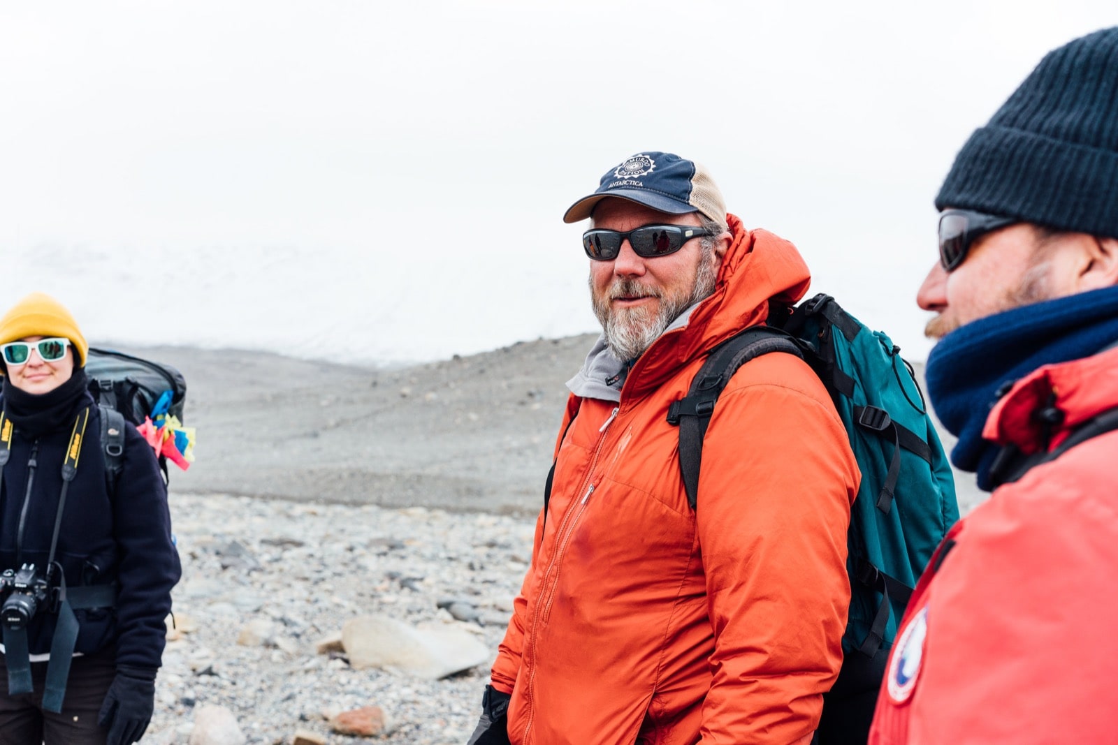  two men with sunglasses and a woman in a yellow beanie and sunglasses smile in a gray rocky landscape. 