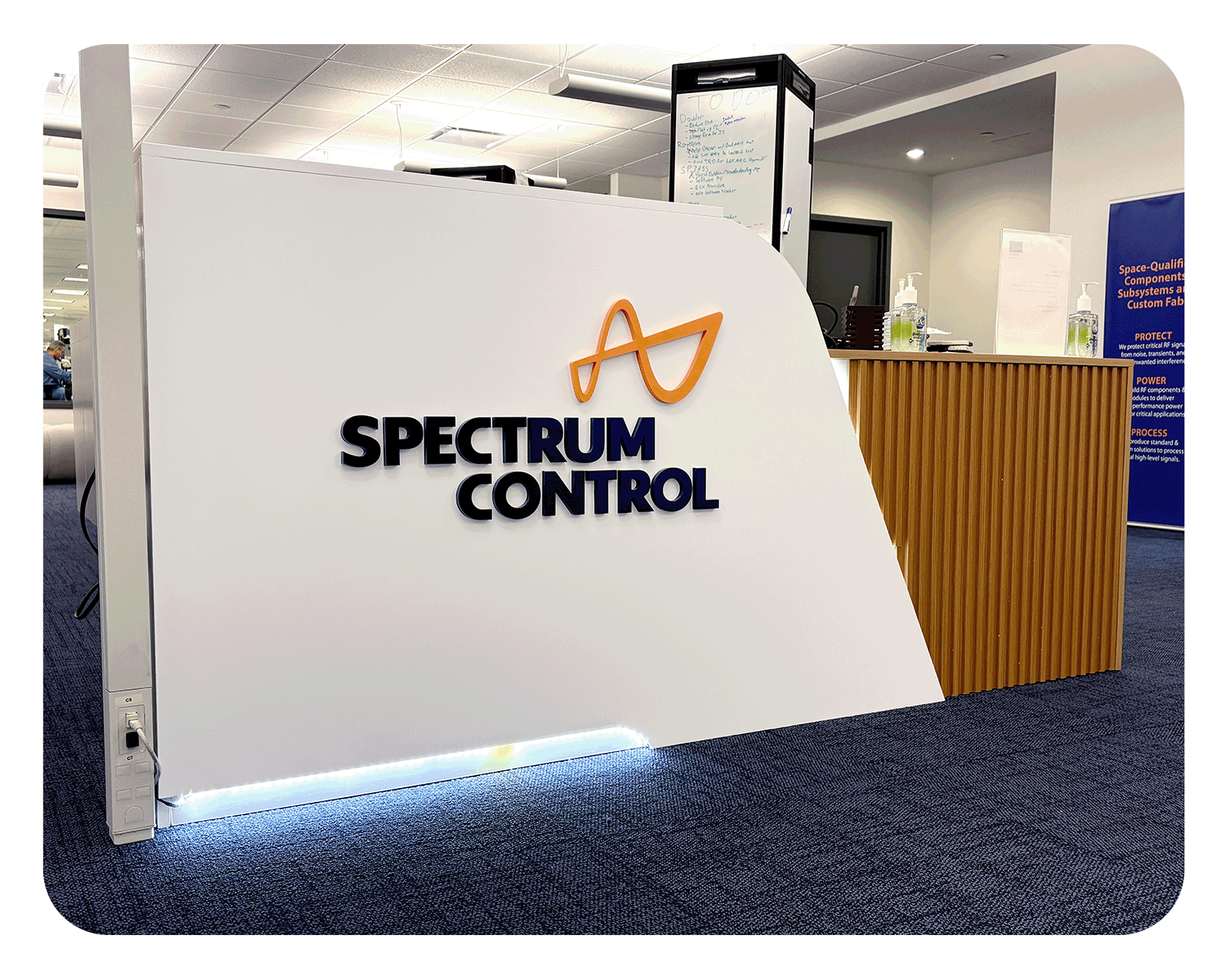 A white reception desk with the Spectrum Control company logo and name displayed on it, situated in an office setting with hand sanitizer bottles on the side and a wooden counter in the background.