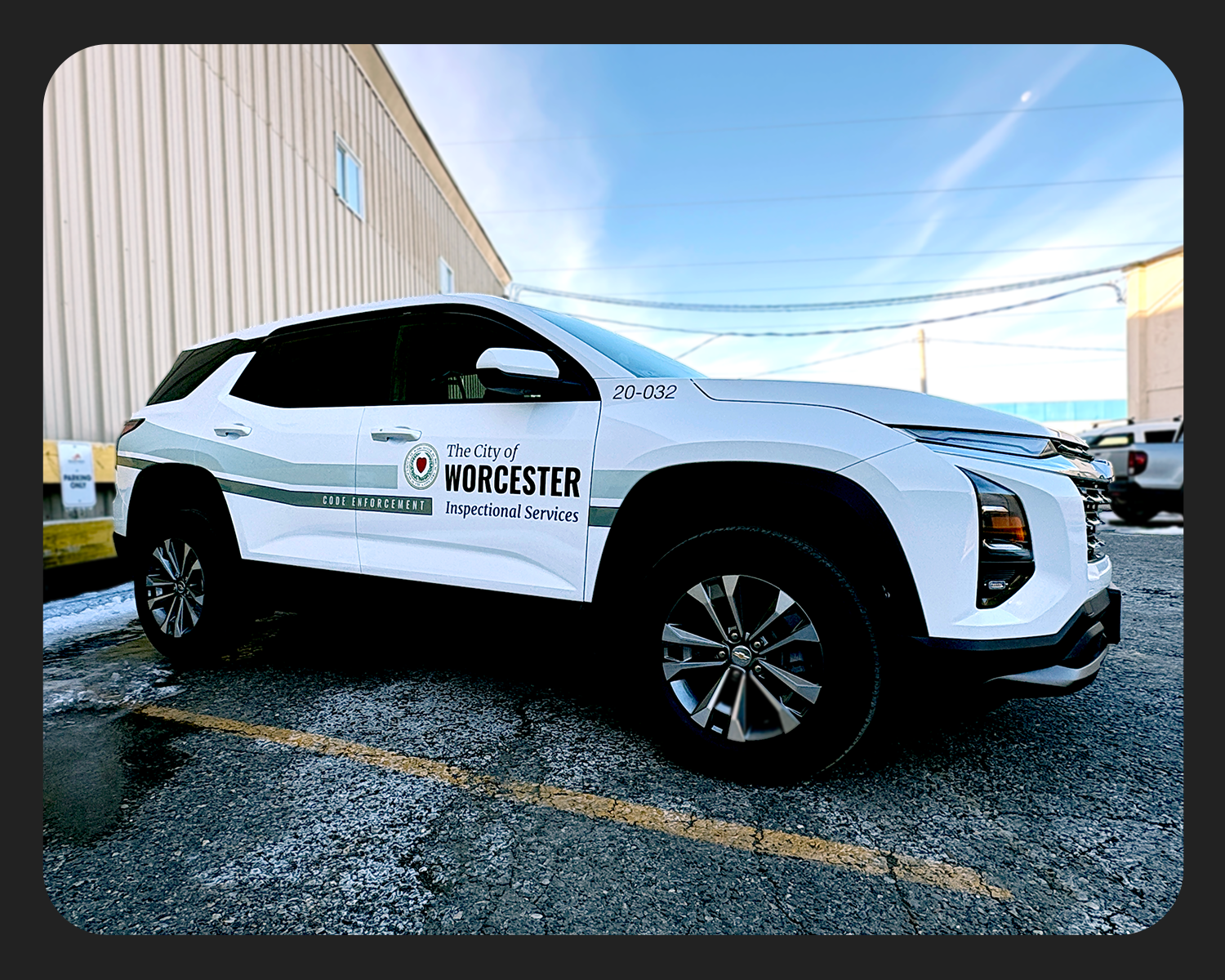 White police vehicle with Worcester city inspection services logo and code enforcement label parked on a wet asphalt lot near a beige industrial building, under a blue sky.