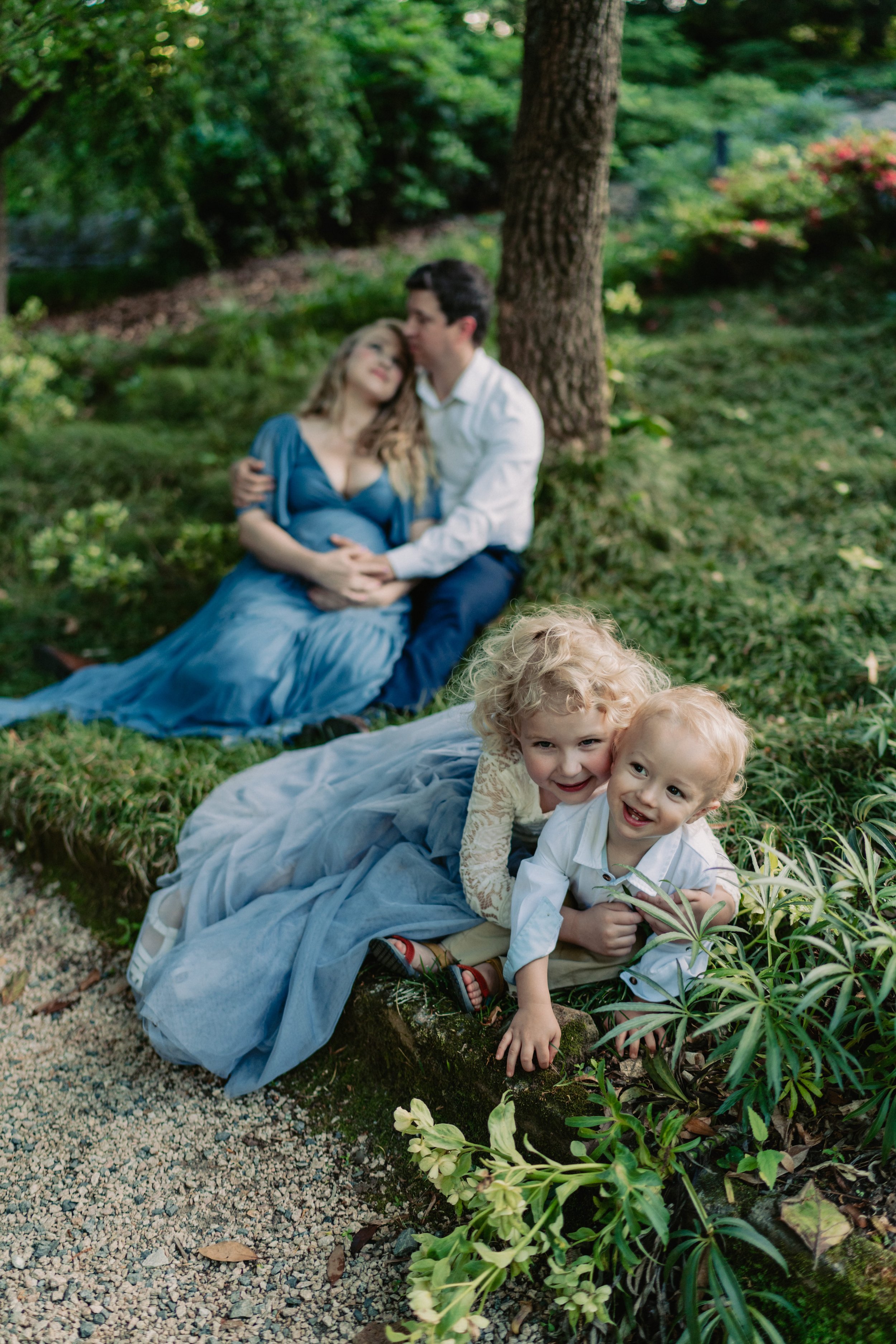 A family with two children lying on a blanket outdoors in a garden, with trees and lush greenery around, enjoying a sunny day.