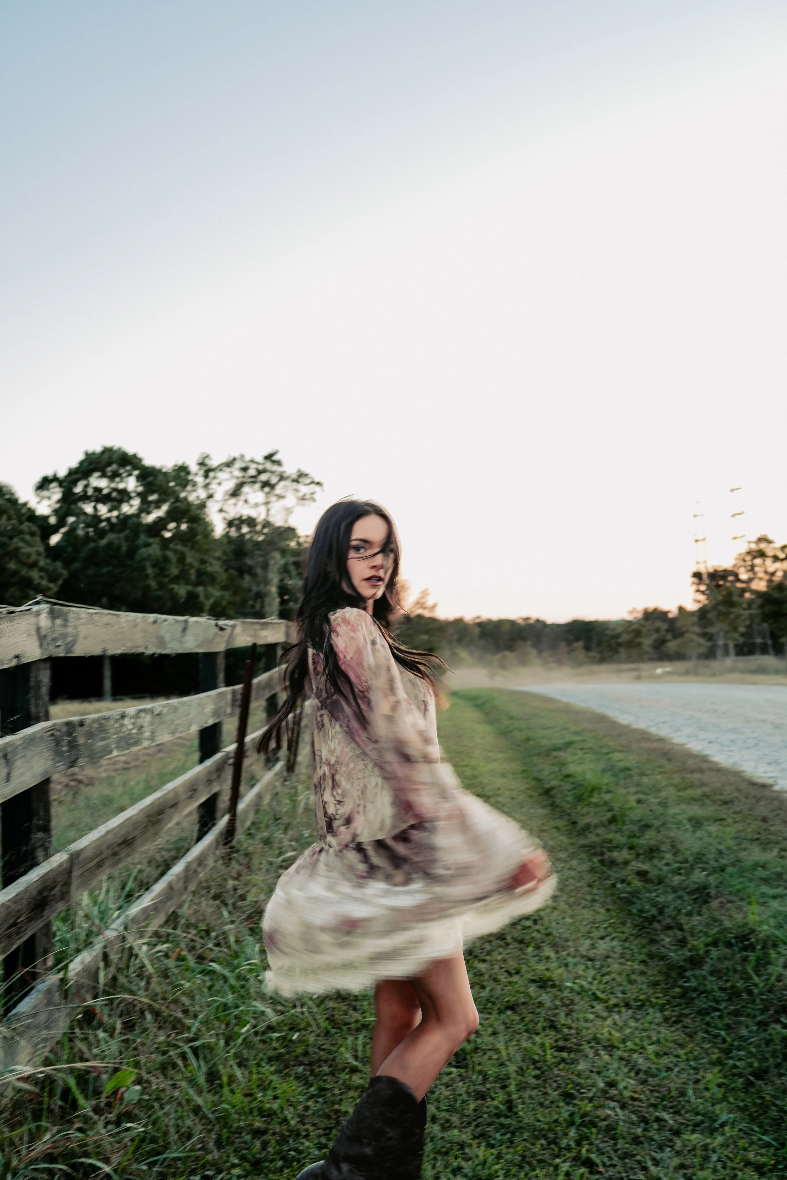 A woman in a flowing floral dress twirls in an open grassy field at sunset, with trees and a wooden fence on the left and power lines in the background.