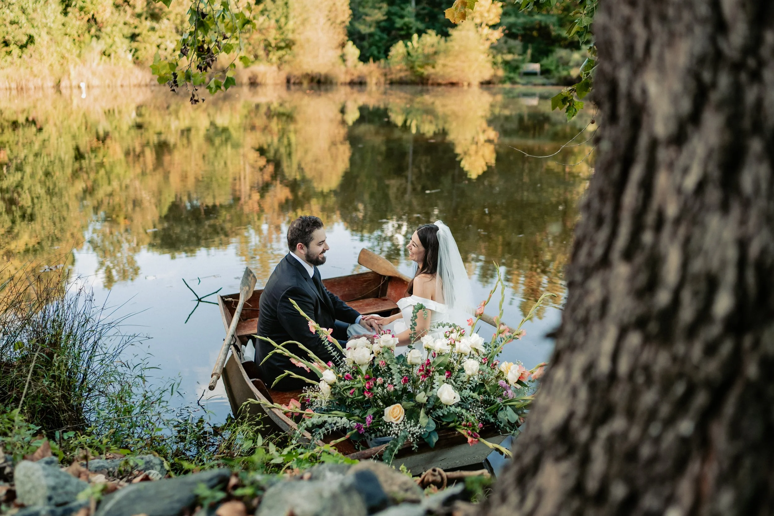 A bride and groom sitting in a small boat on a lake, surrounded by trees with autumn foliage, holding hands and looking at each other, with a large bouquet of flowers in the boat.