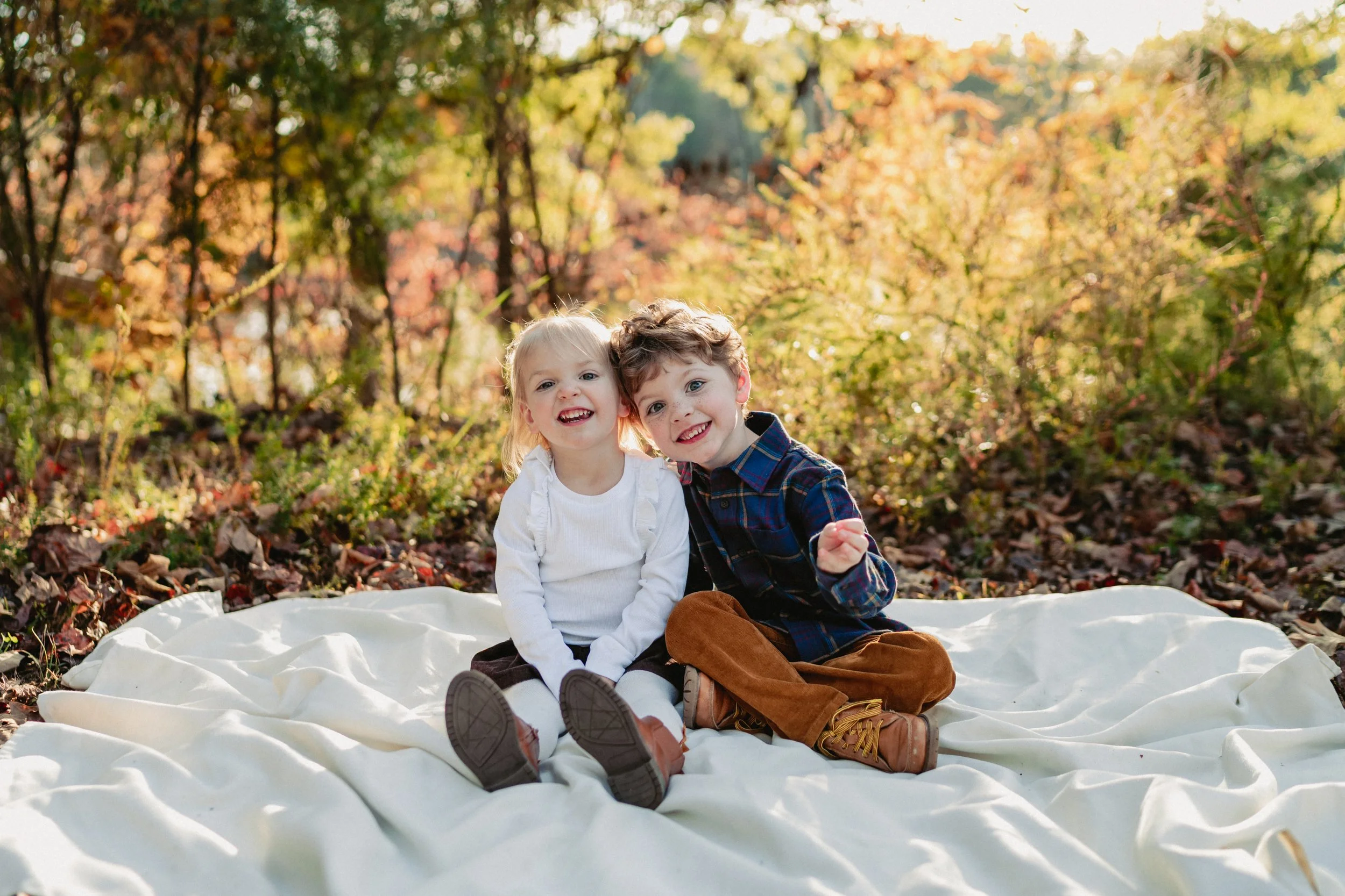 Two young children, a girl and a boy, sitting together on a white blanket outdoors surrounded by autumn leaves with trees showing fall foliage in the background, smiling and enjoying a sunny day.
