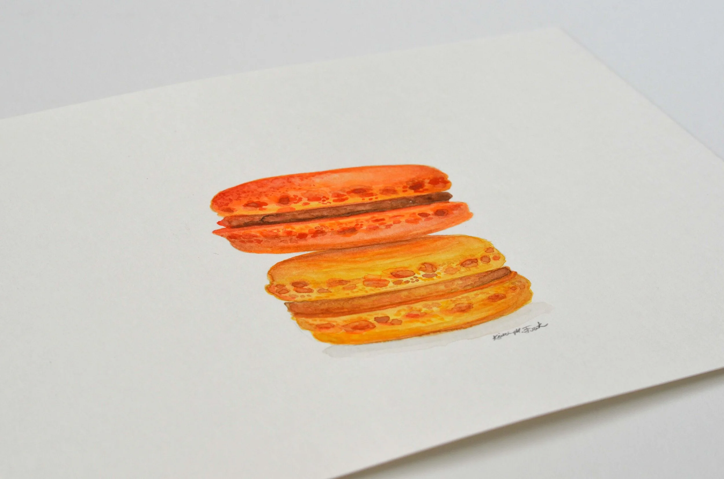 Photo of the watercolor titled "Macaron Monday No.1" by Kristin Frank. The painting features a detailed rendering of two macarons stacked on top of each other. One is orange and one is yellow.