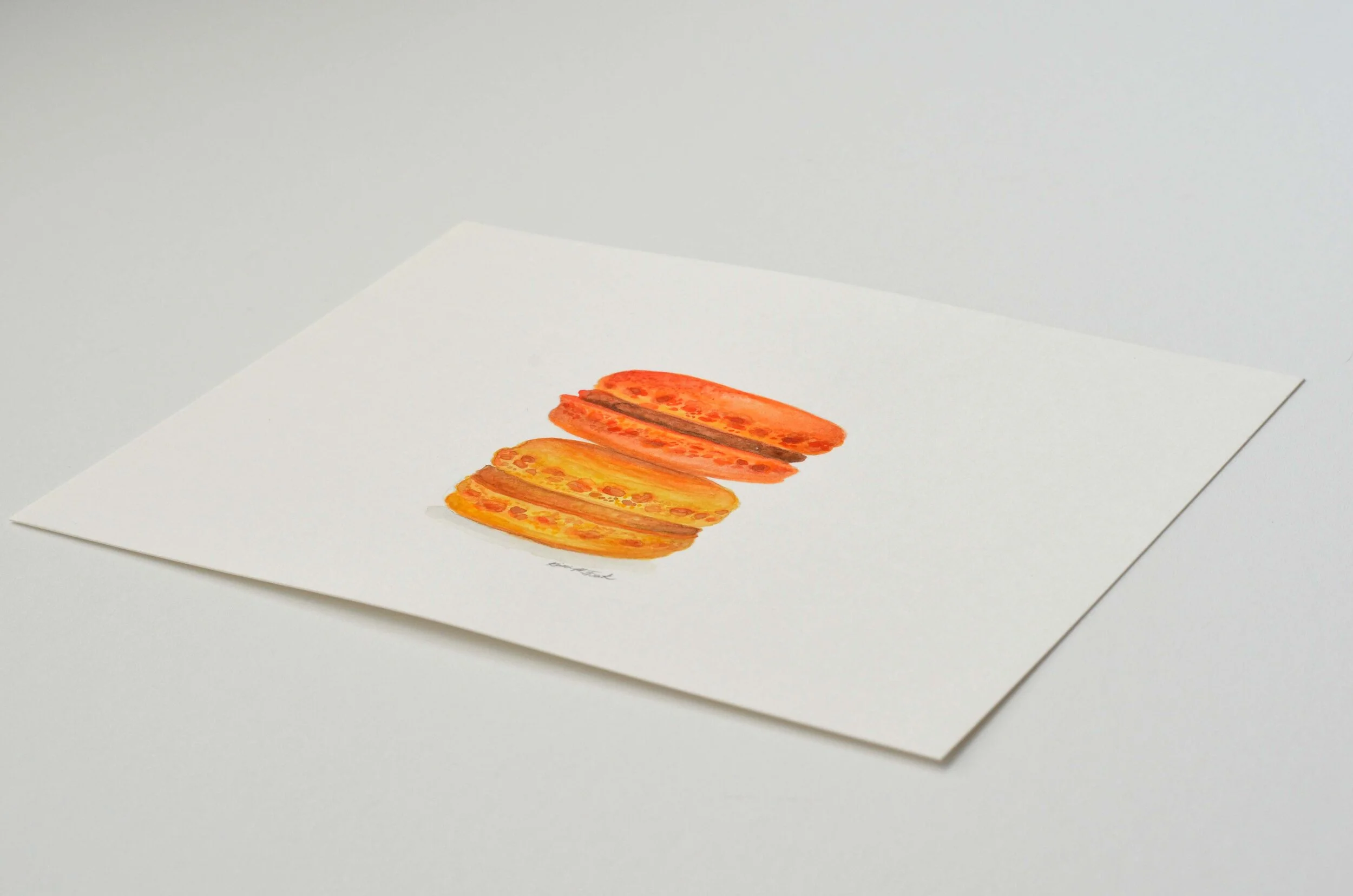 Photo of the watercolor titled "Macaron Monday No.1" by Kristin Frank. The painting features a detailed rendering of two macarons stacked on top of each other. One is orange and one is yellow.