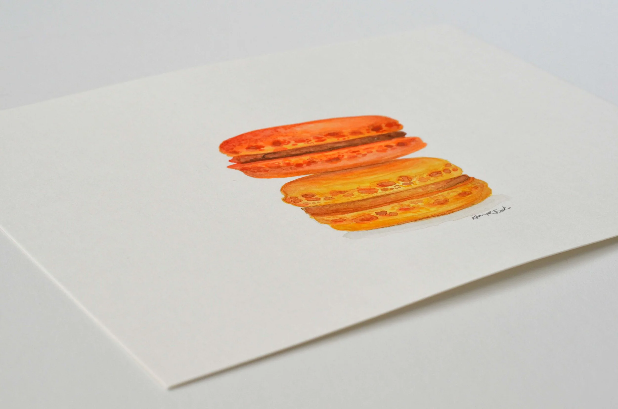 Photo of the watercolor titled "Macaron Monday No.1" by Kristin Frank. The painting features a detailed rendering of two macarons stacked on top of each other. One is orange and one is yellow.